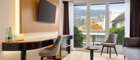 Ein modernes Hotelzimmer mit einem wandmontierten Flachbildfernseher, einem Holztisch mit Stuhl, einem runden Tisch mit Stuhl, einer Couch und einem großen Fenster mit Blick auf einen Balkon und die umliegende Landschaft.