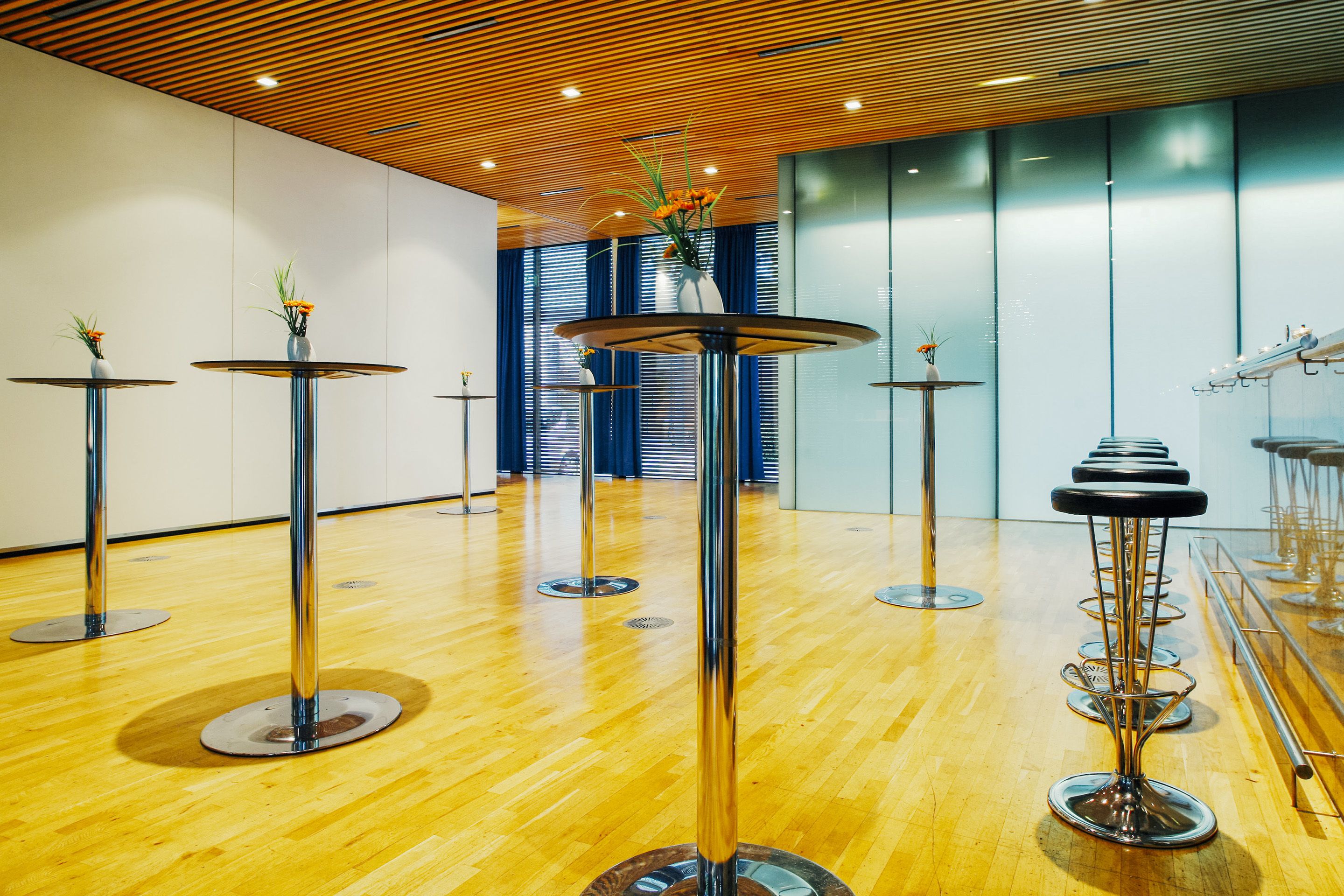 A modern interior with multiple round tables and chairs, wooden flooring, and a ceiling with wooden slats.
