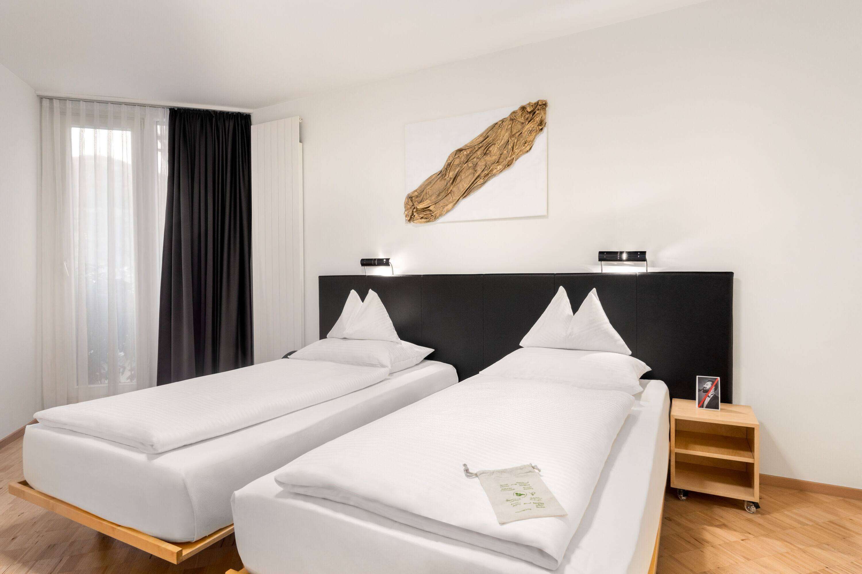 Habitación de hotel con dos camas individuales, una mesita de noche de madera, un cuadro enmarcado en la pared y una ventana con cortinas.