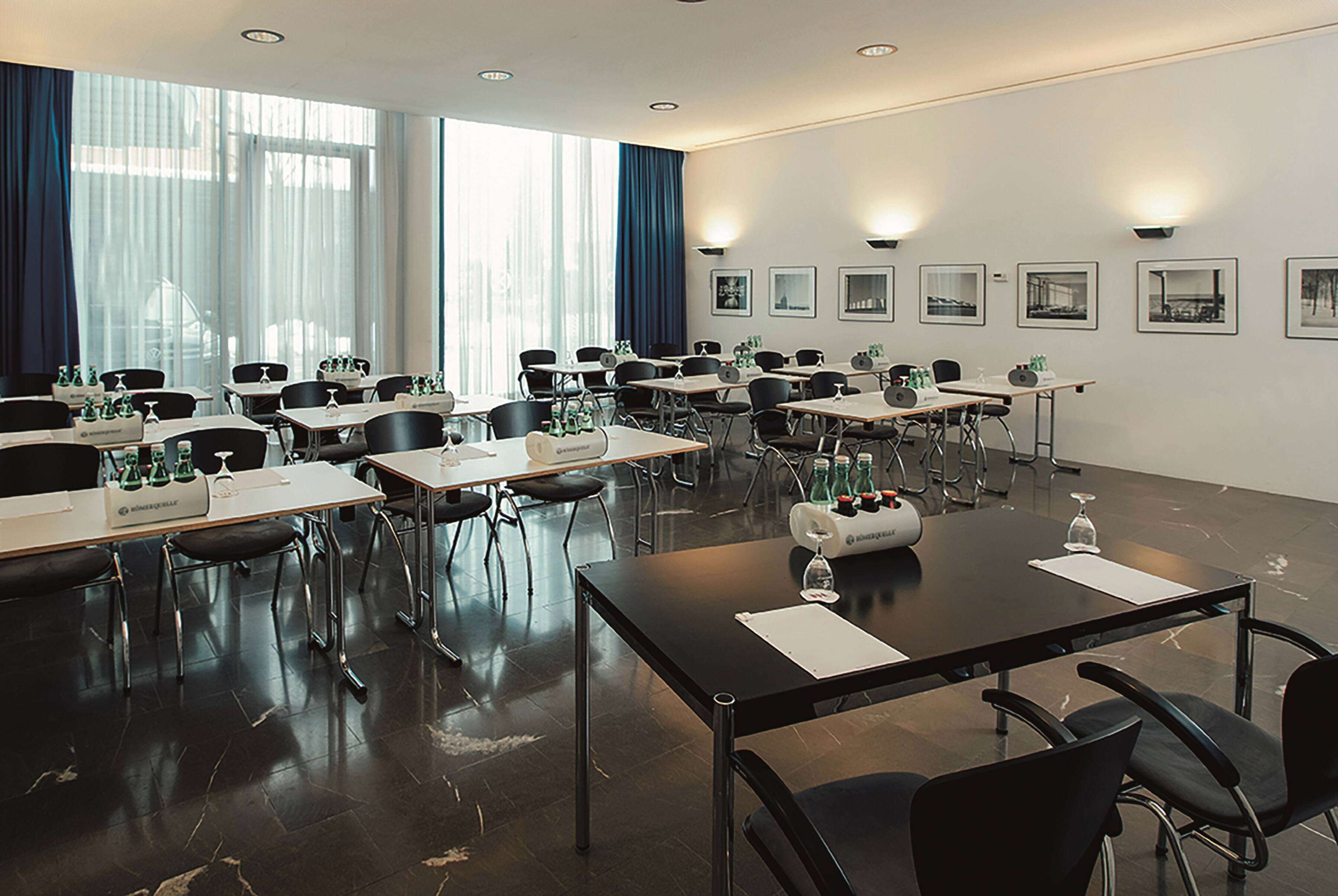 Ein Konferenzraum mit mehreren Tischen und Stühlen, großen Fenstern mit Vorhängen und gerahmten Bildern an den Wänden.