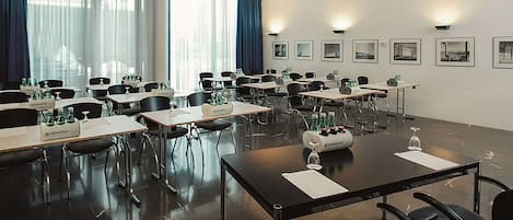 Un salón de conferencias con varias mesas y sillas, ventanas amplias con cortinas, y cuadros en las paredes.