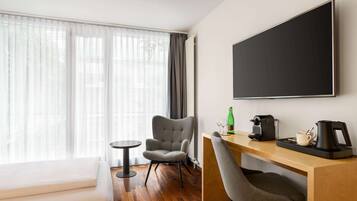 Une chambre d’hôtel moderne avec un bureau en bois, deux chaises, une grande fenêtre avec des rideaux légers, un téléviseur à écran plat et une machine à café.