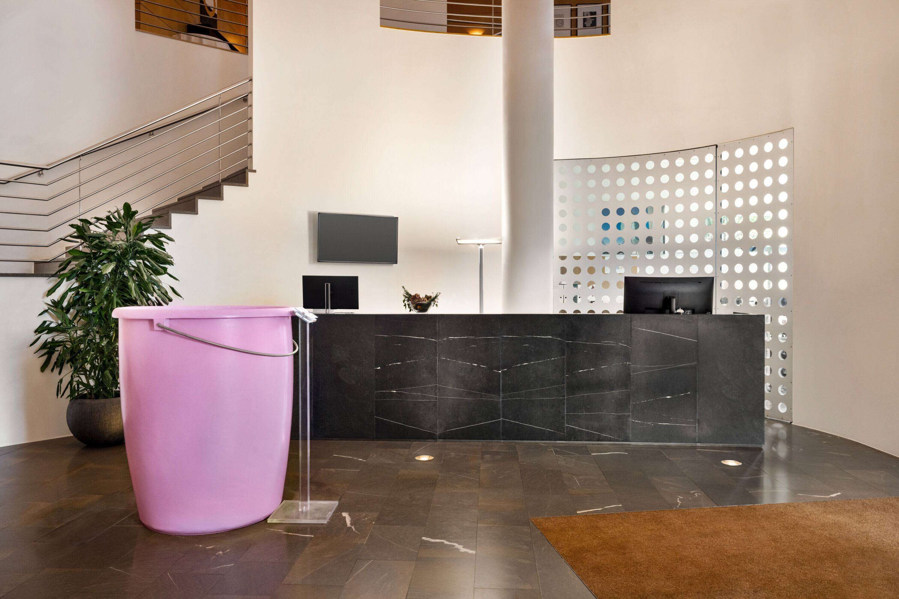 Zona de recepción con mostrador de mármol negro, un cubo rosa, una planta en maceta y una escalera al fondo.