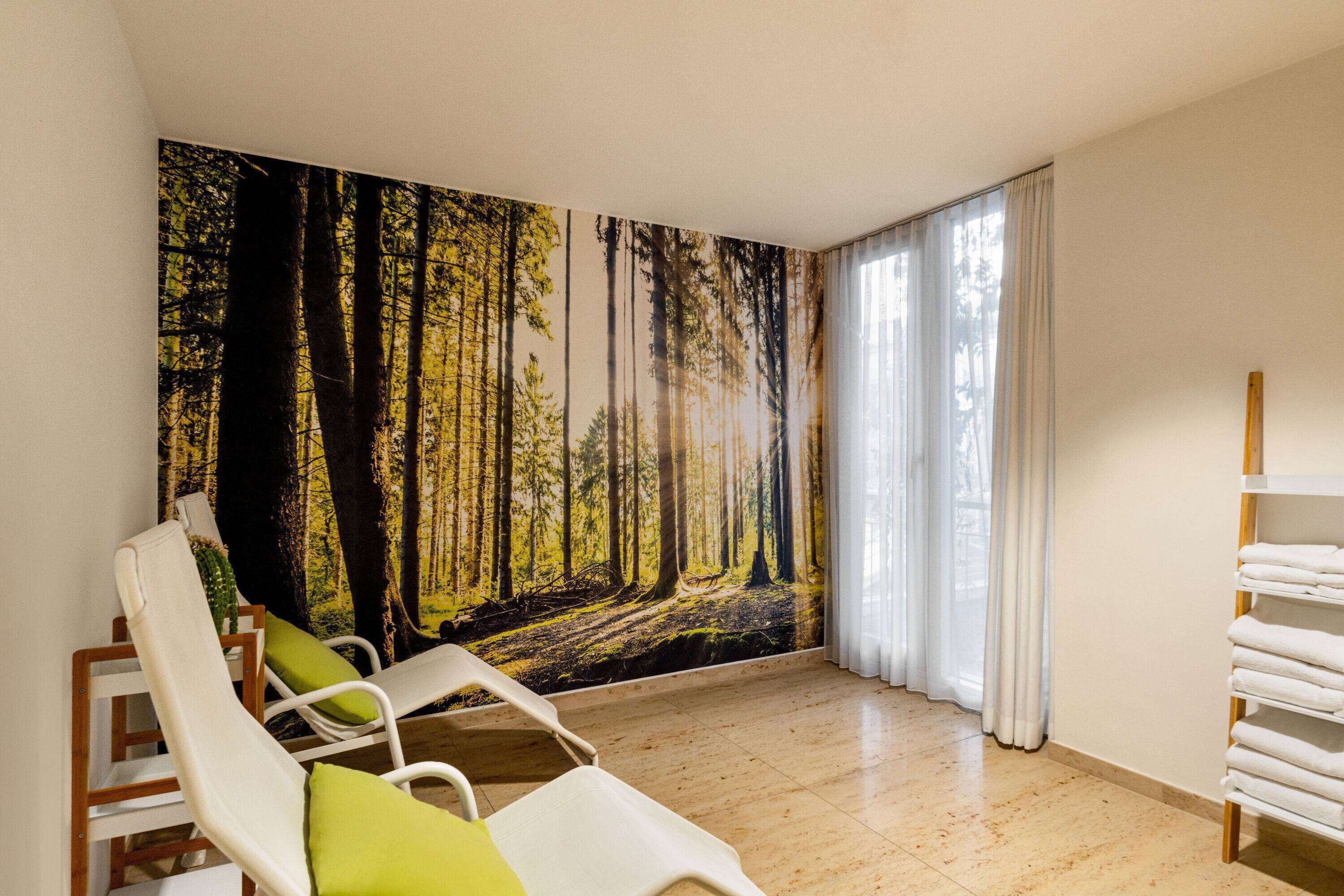 A room with two white lounge chairs, a large forest mural on the wall, a wooden shelf with towels, and a window with white curtains.