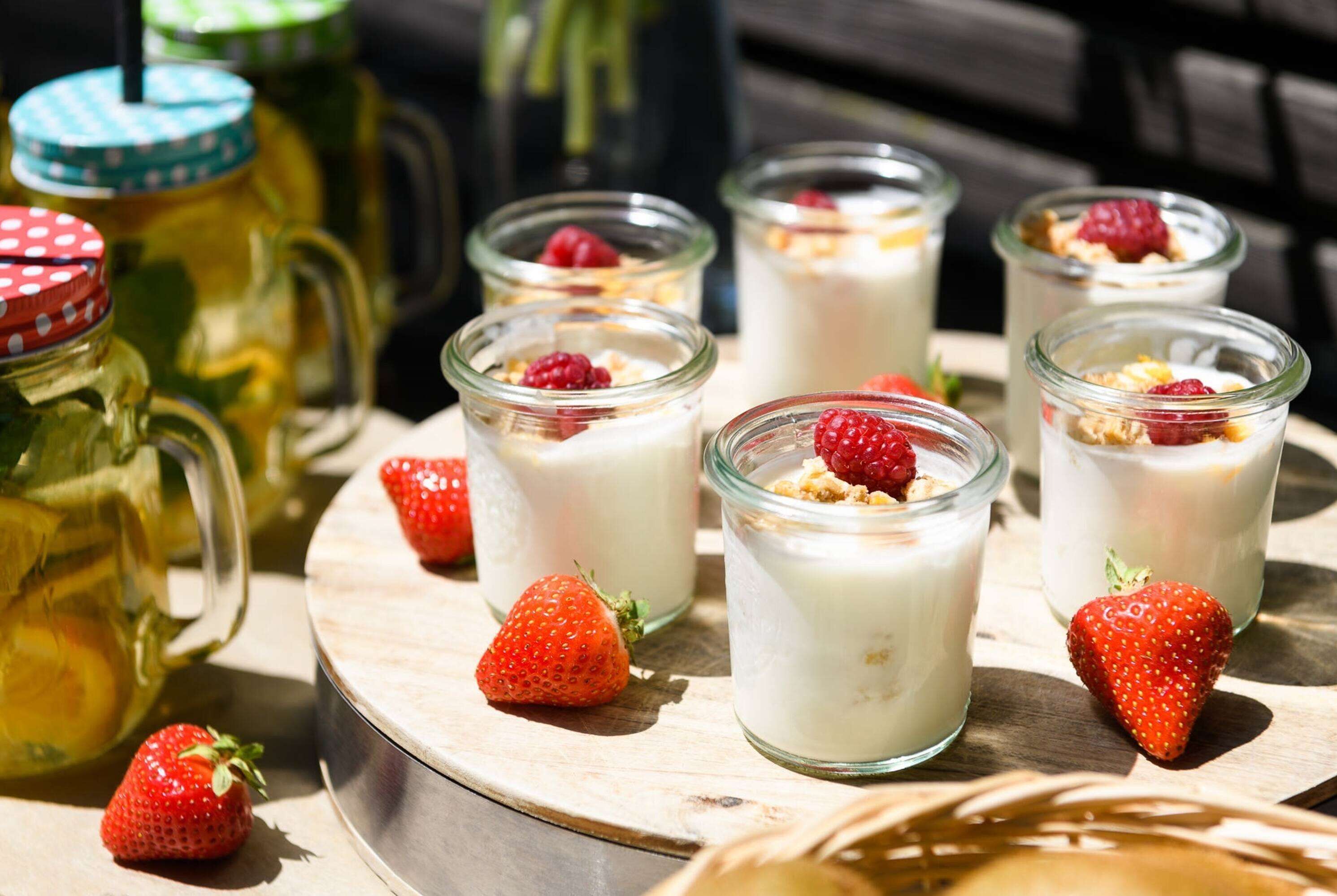 Six glass jars of yogurt topped with granola and berries, a pitcher of lemonade, and fresh strawberries on a wooden tray.