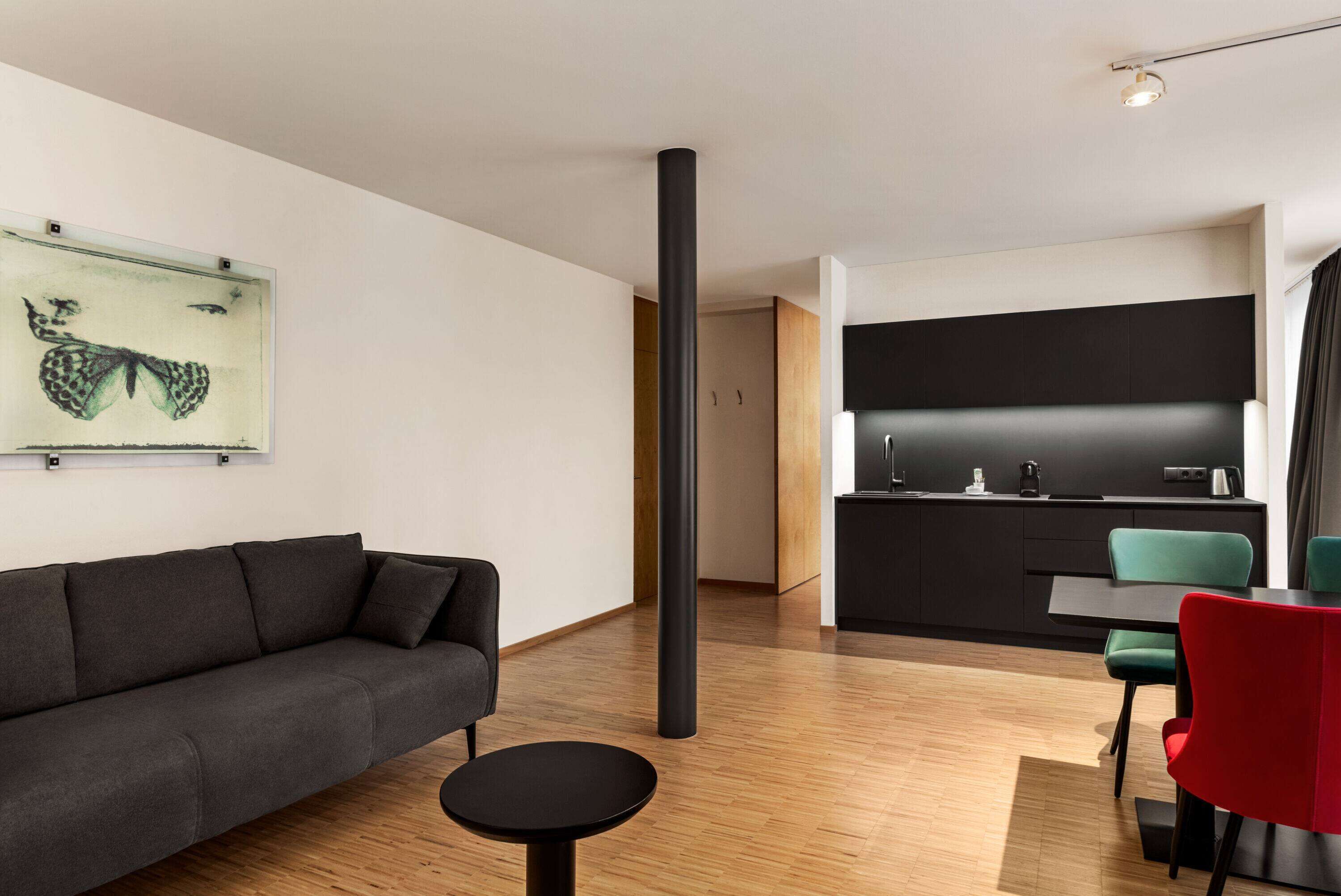 A modern living room with a sofa, a small round table, and a kitchen area with a black countertop and green chairs.