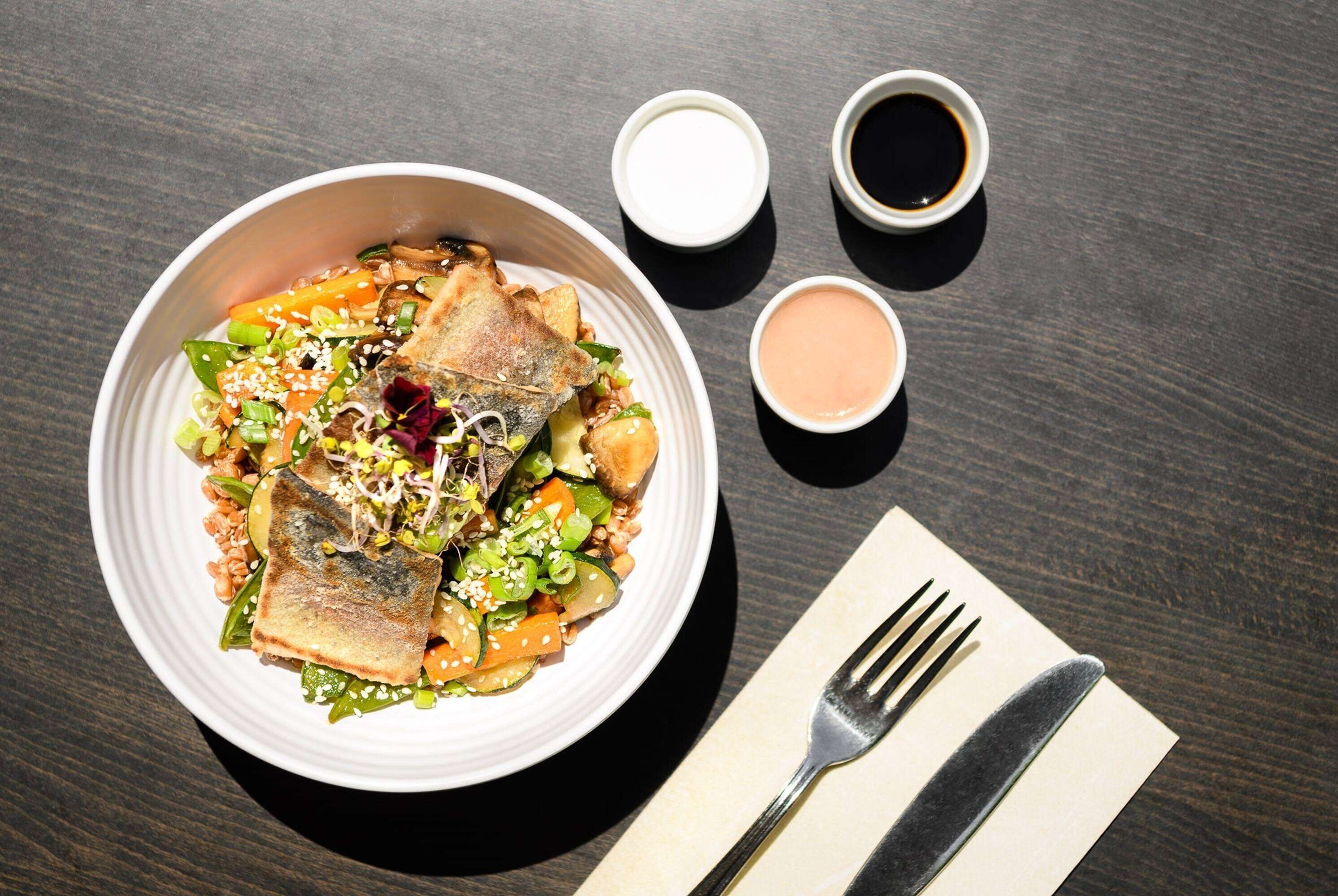 A bowl of food with a fork and knife on a napkin, and two small bowls of sauce on a table.