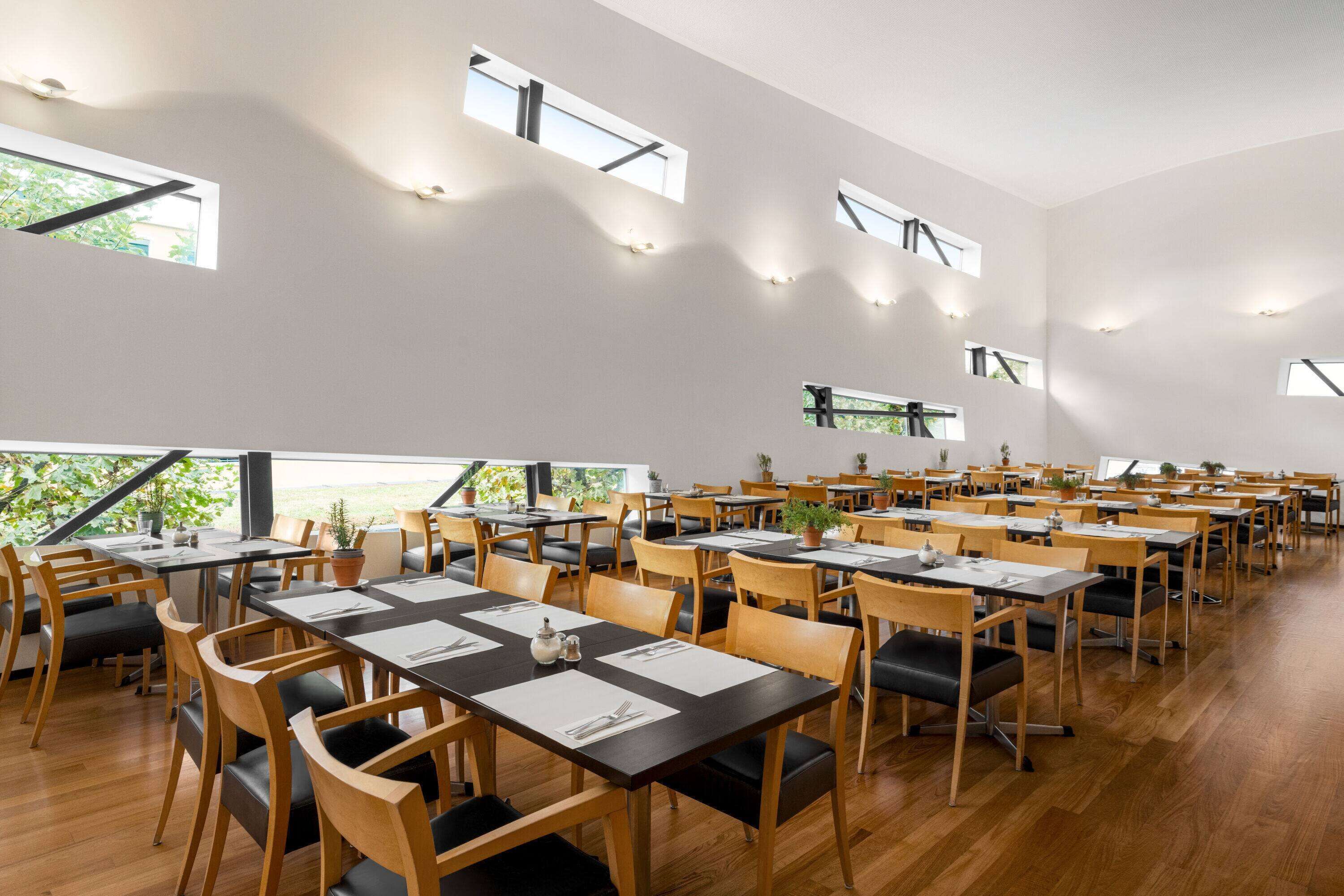 A spacious dining area with multiple tables and chairs, large windows, and a view of greenery outside.
