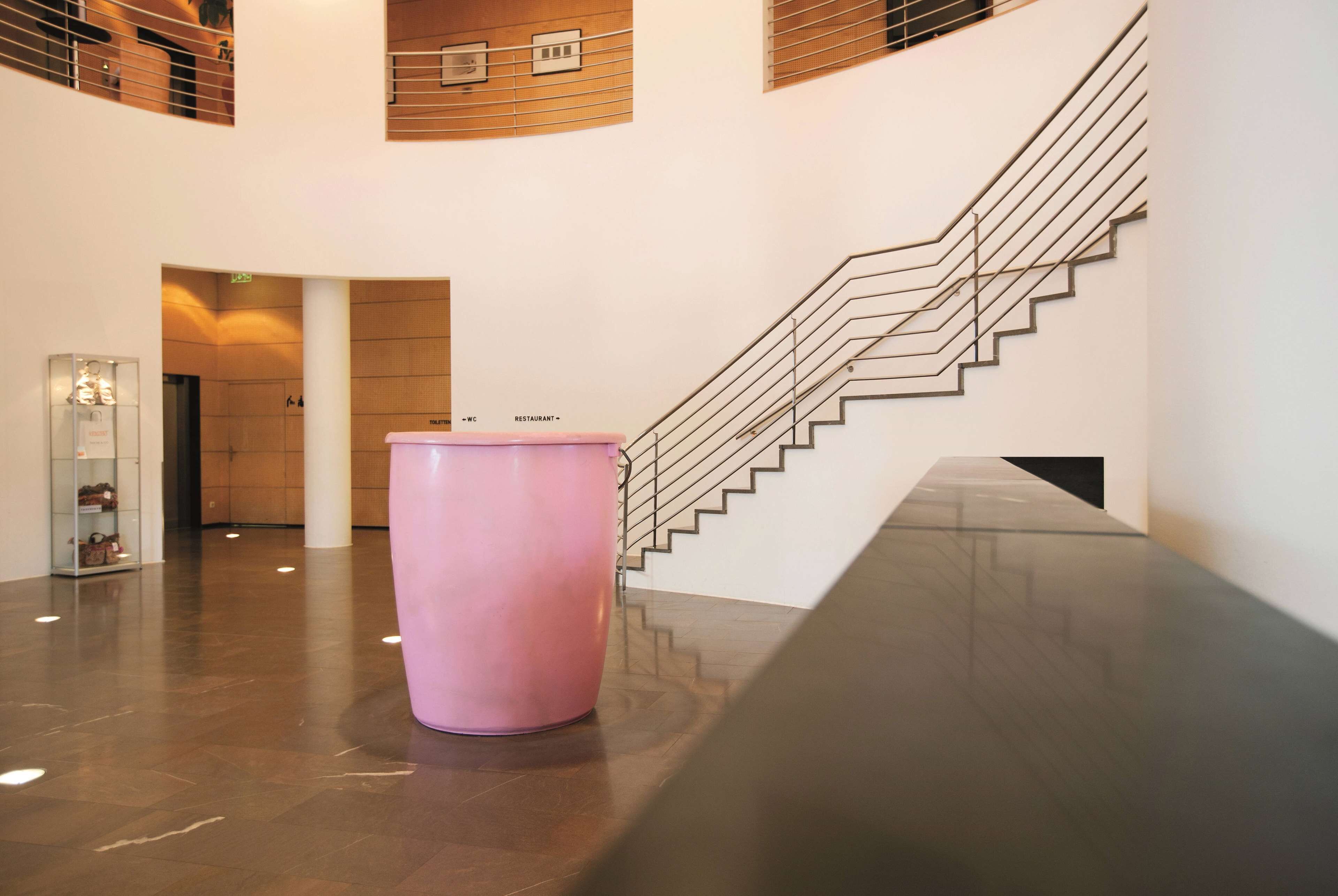 Een moderne lobby met een roze cilindervormig object, een trap en een glazen vitrine.