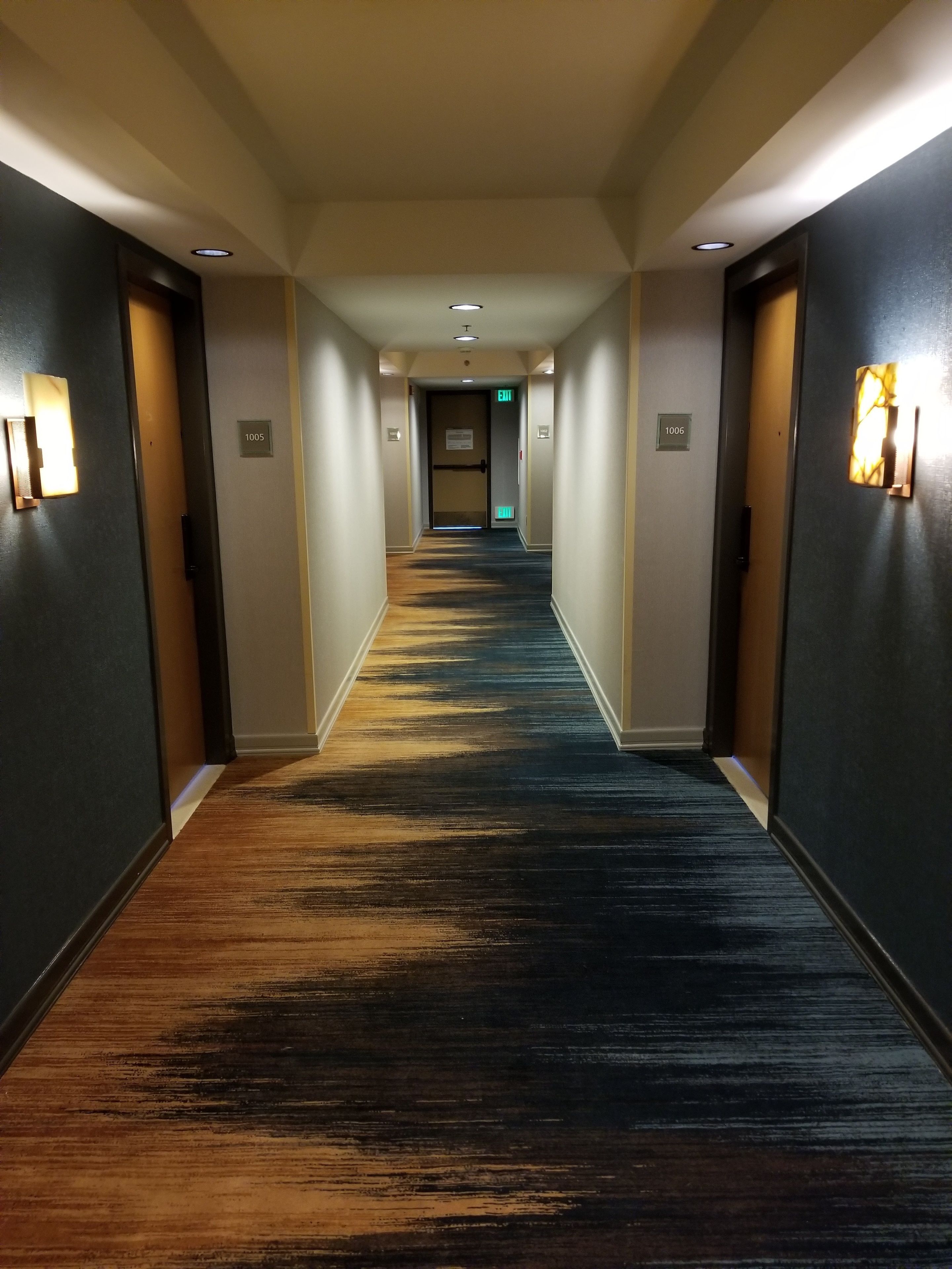 Un pasillo de hotel con piso alfombrado, paredes con luces y puertas con los números 1005 y 1006.