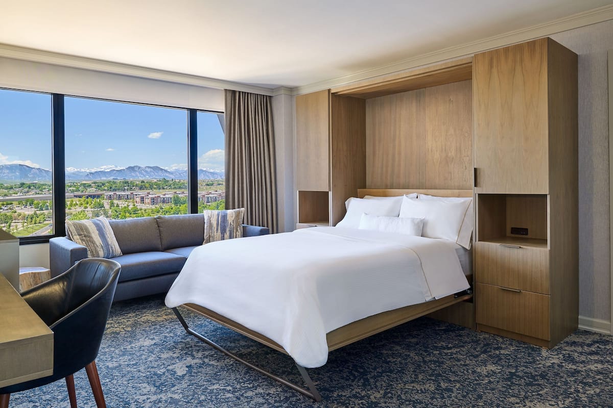 Ein Hotelzimmer mit einem großen Bett, einer Couch, einem Schreibtisch und Blick auf eine Stadtlandschaft und Berge.