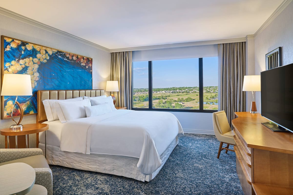 Ein Hotelzimmer mit einem großen Bett, zwei Nachttischen mit Lampen, einem Schreibtisch mit Fernseher und einem großen Fenster mit Blick auf die Landschaft.