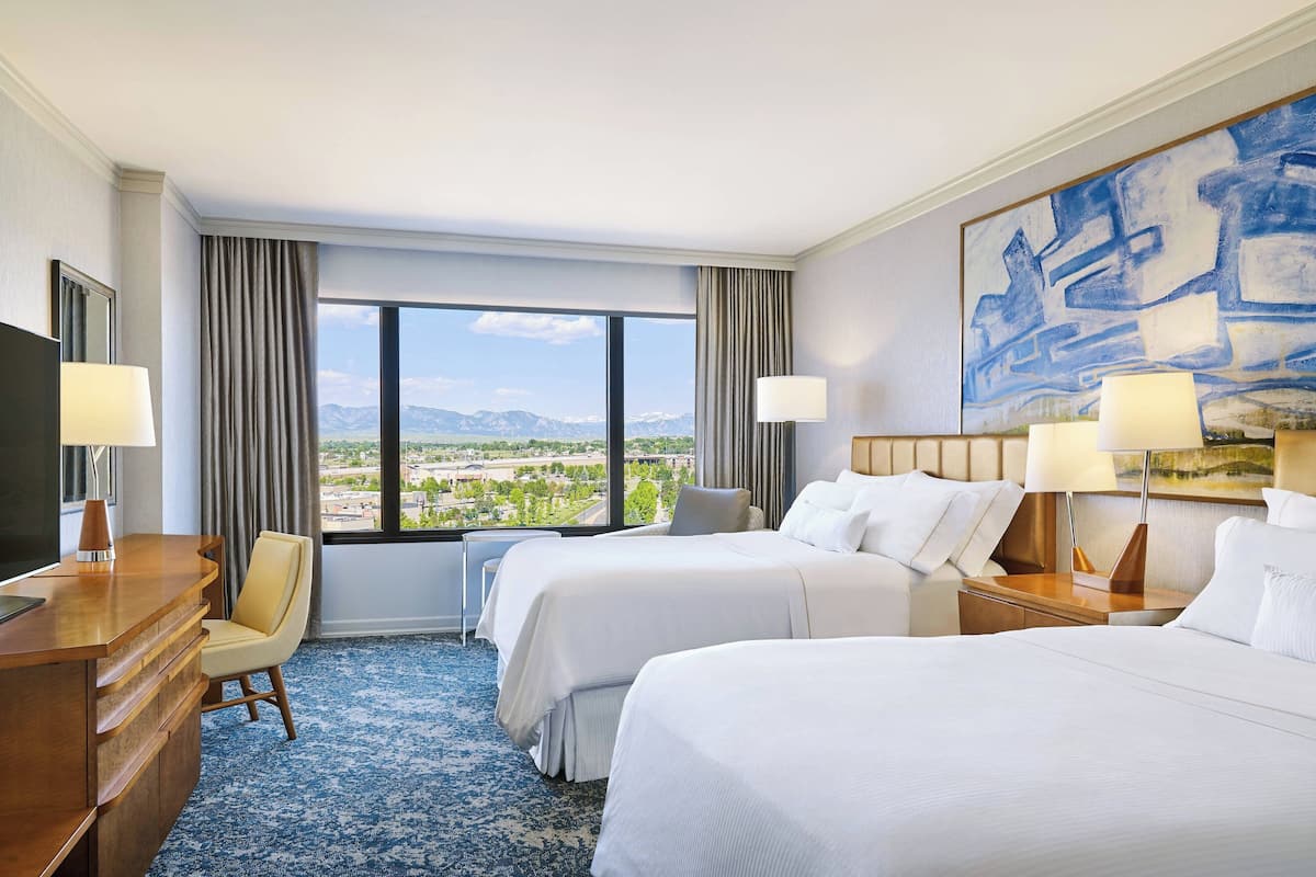 Ein Hotelzimmer mit zwei Betten, einem Schreibtisch, einem Sessel und einem großen Fenster mit Blick auf die Berge.