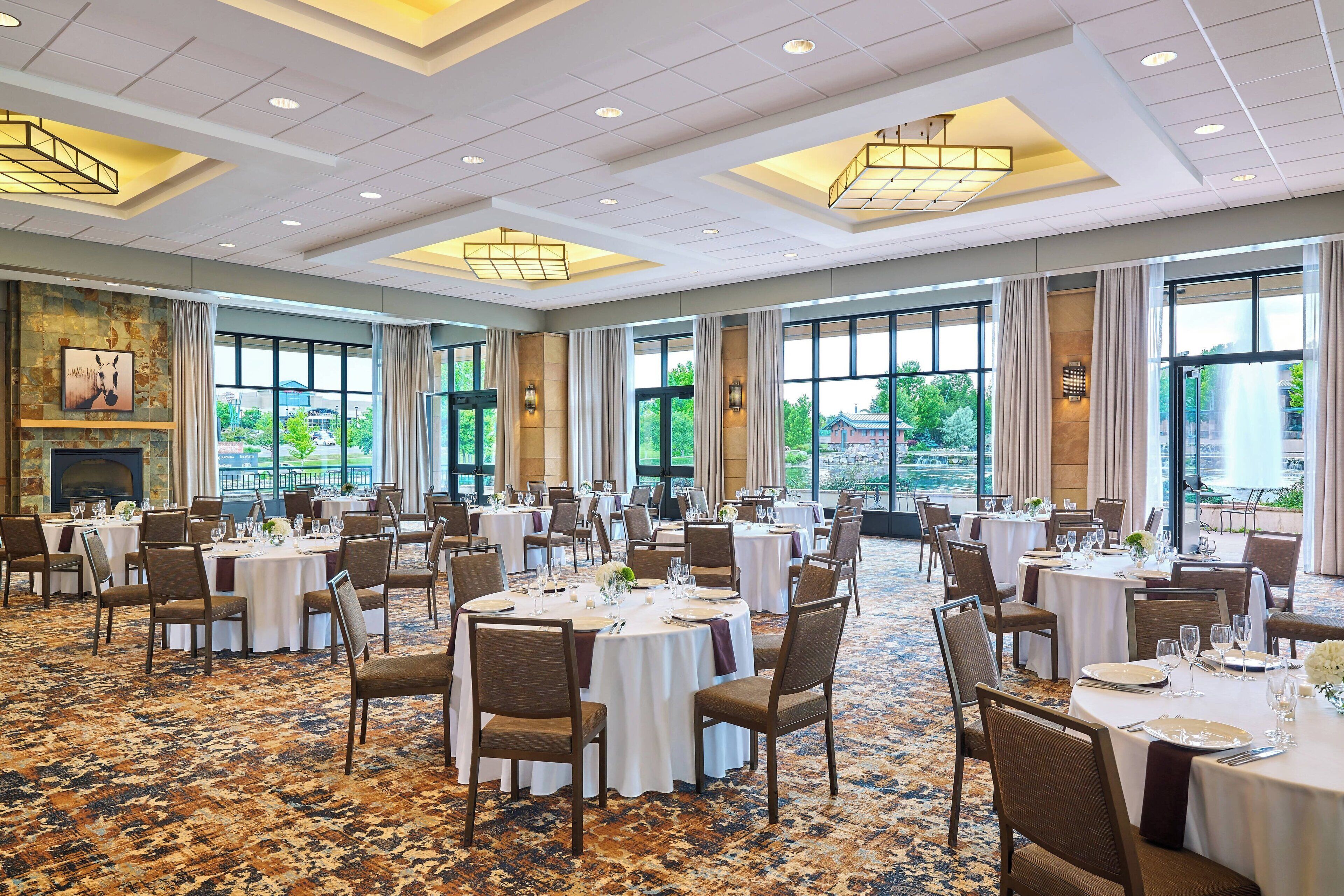 A spacious banquet hall with multiple round tables set for dining, large windows offering a view of greenery outside, and a modern ceiling design with recessed lighting.
