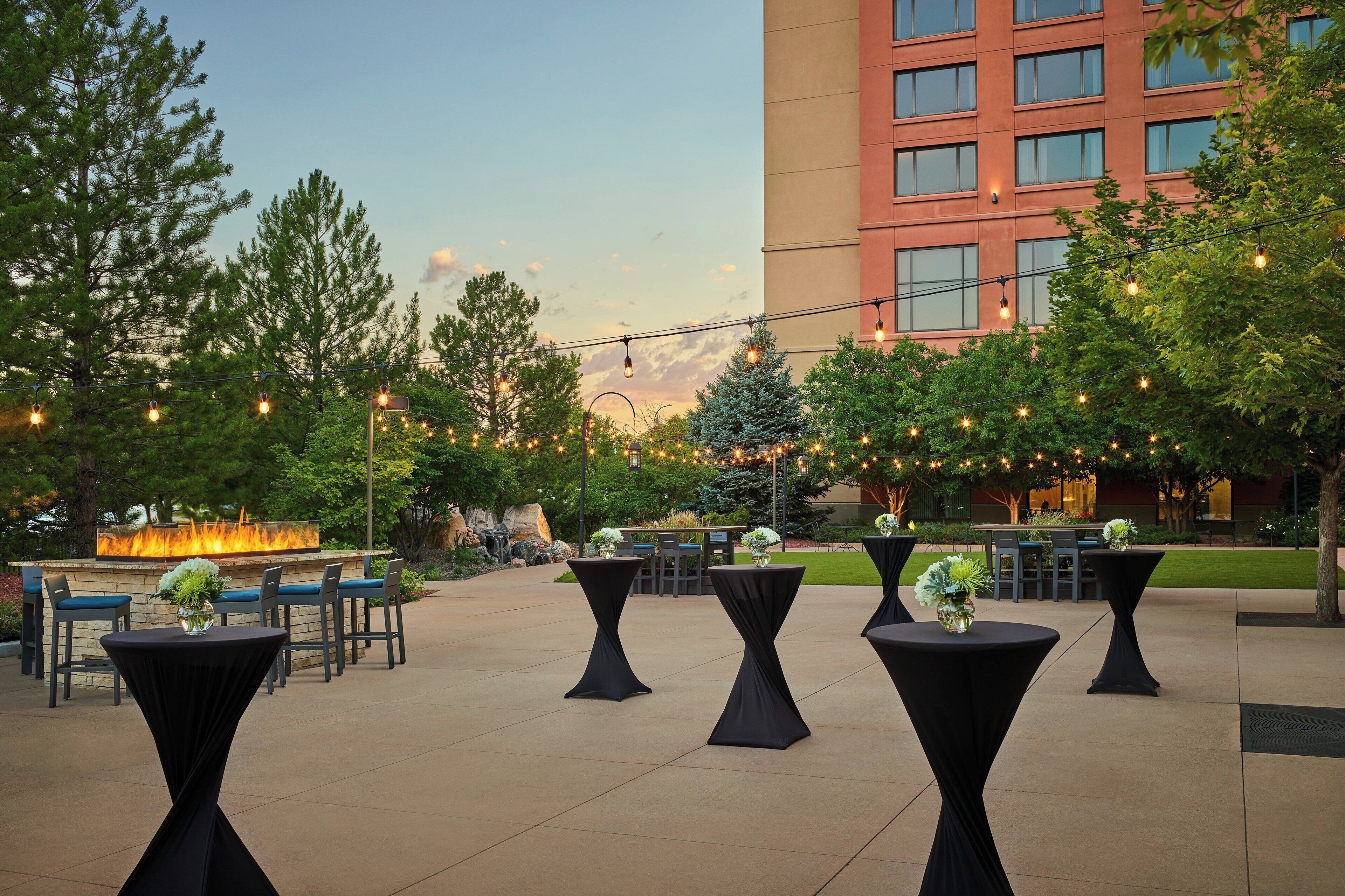 An outdoor patio area with round tables and chairs, string lights, and a fire feature in the background.