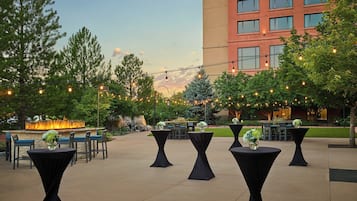 An outdoor patio area with round tables and chairs, string lights, and a fire feature in the background.