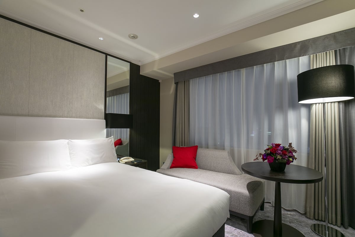 A hotel room with a large bed, a sofa, a small table with a vase of flowers, a lamp, and a large mirror.