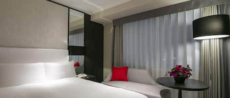 Une chambre d’hôtel avec un grand lit, un canapé, une petite table sur laquelle est posé un vase de fleurs, une lampe et un grand miroir.