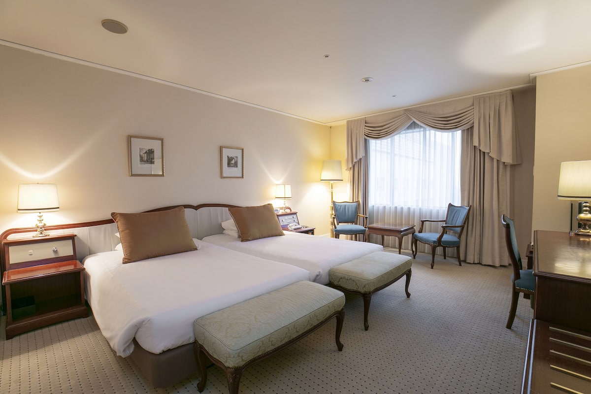 A hotel room with two beds, a desk, a chair, and a lamp.