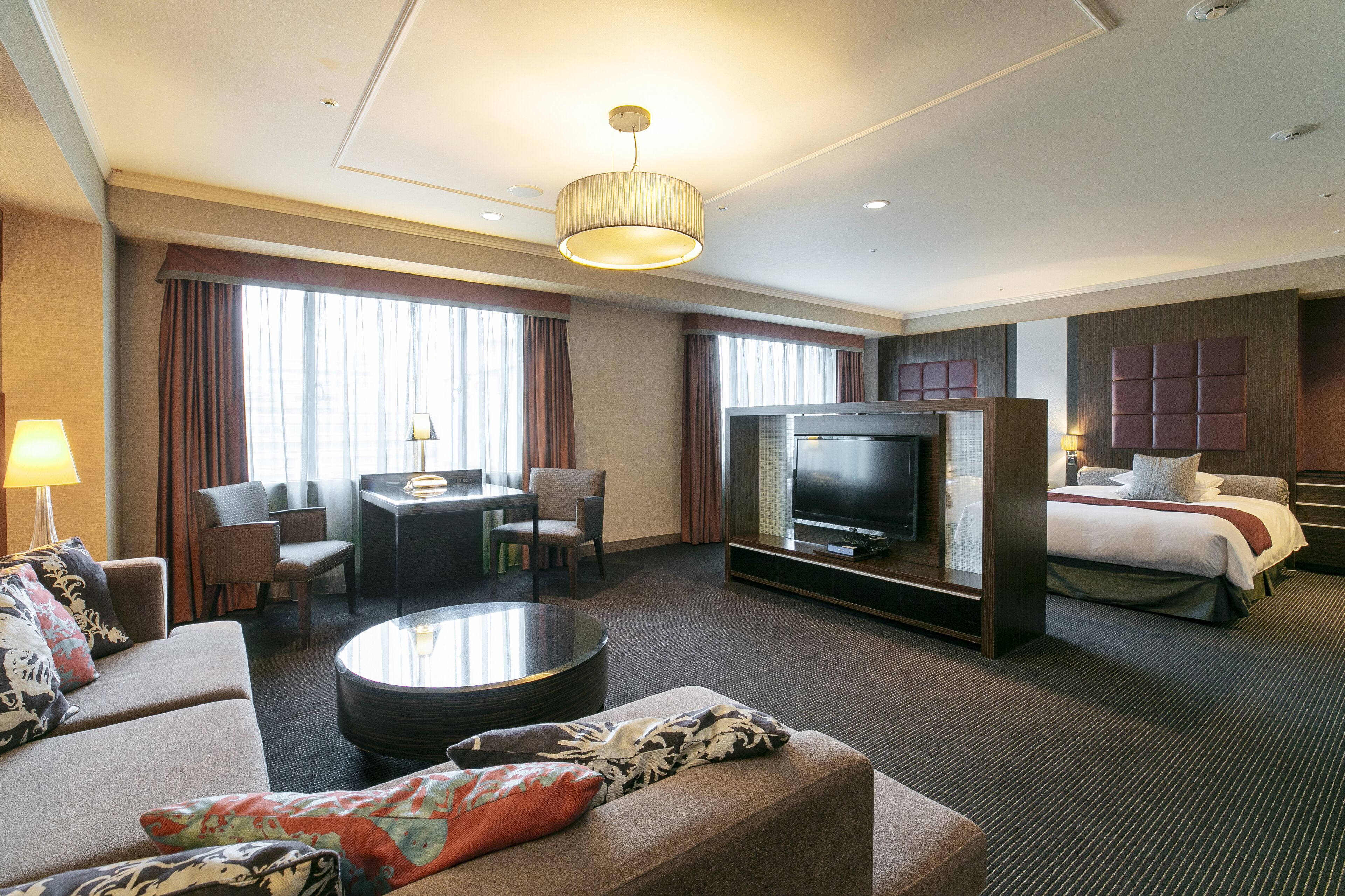A hotel room with a sofa, a coffee table, a TV, a bed, and a desk with chairs.