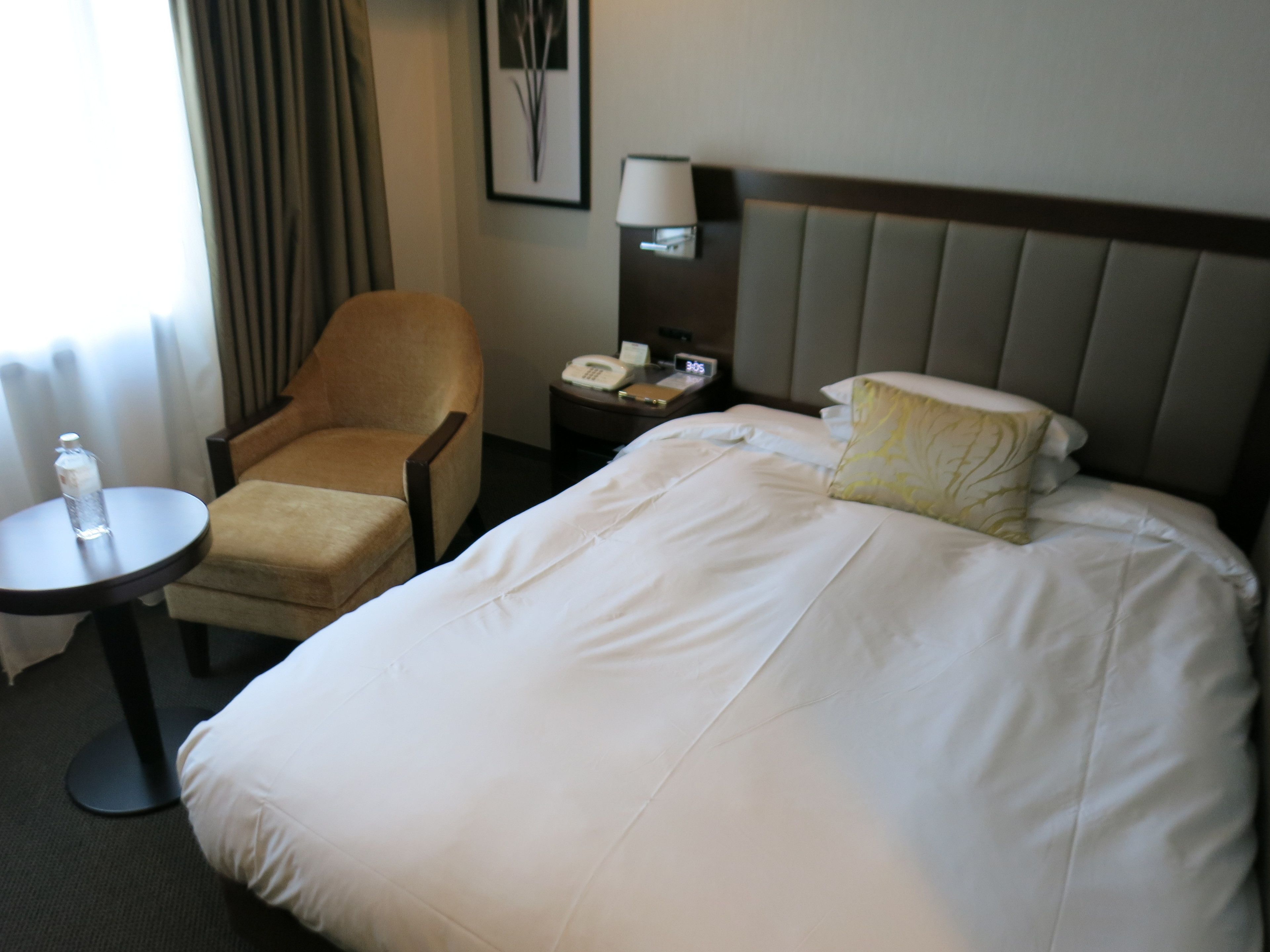 A hotel room with a bed, a chair, a nightstand with a phone, and a lamp.