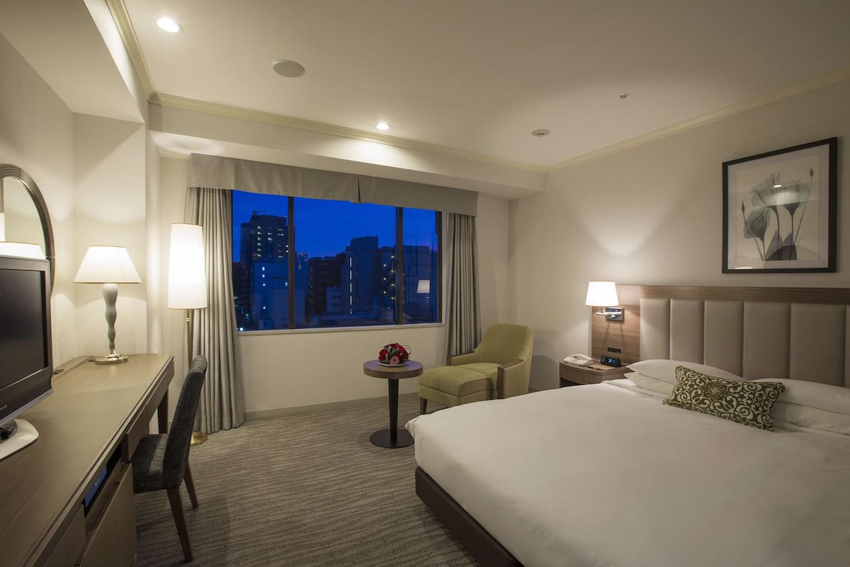 A hotel room with a bed, a desk with a chair, a small table with a lamp, a nightstand with a lamp, and a window with a view of the city at dusk.