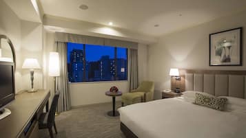 A hotel room with a bed, a desk with a chair, a small table with a lamp, a nightstand with a lamp, and a window with a view of the city at dusk.