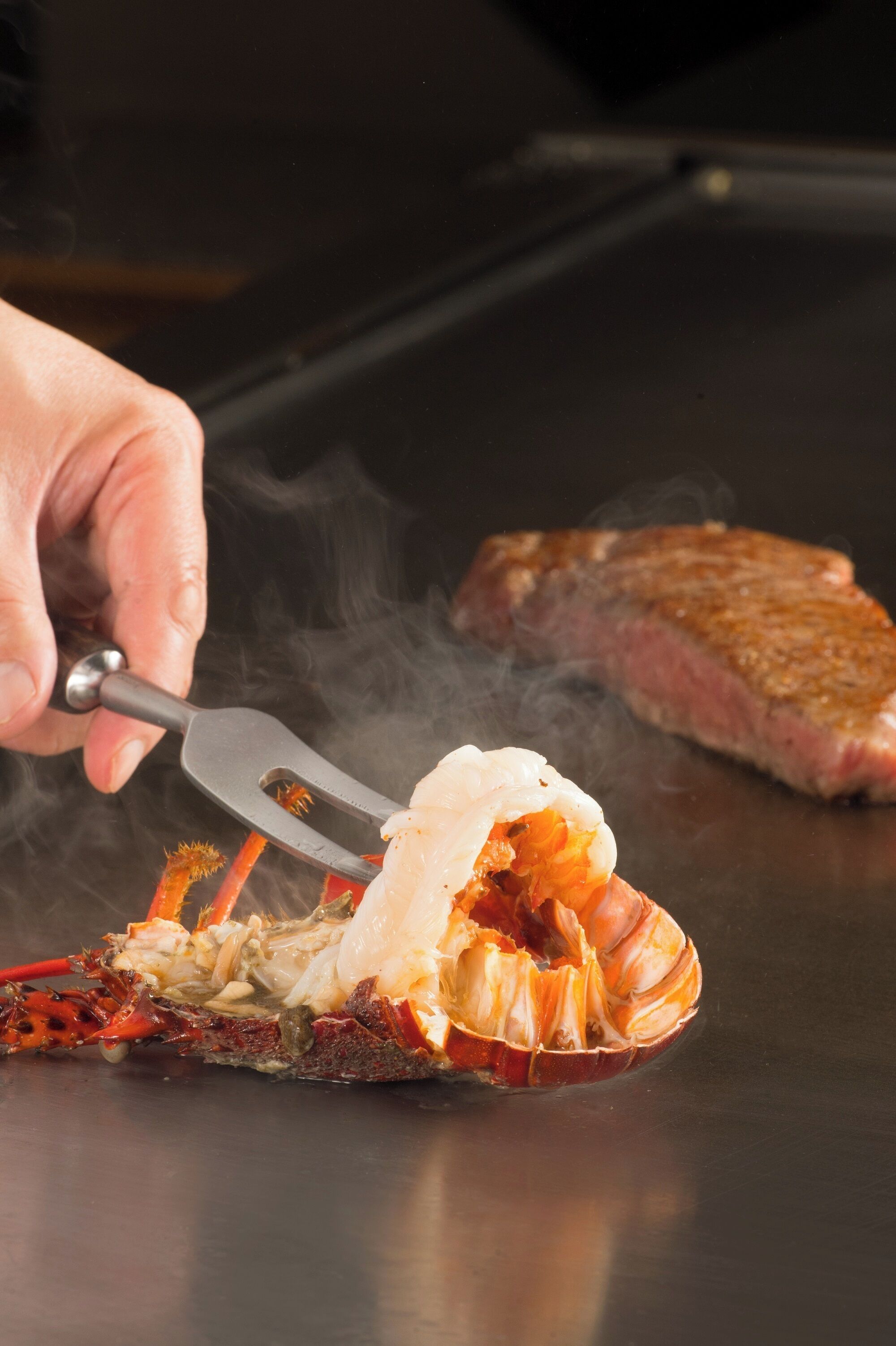 A person using tongs to handle a lobster on a grill.