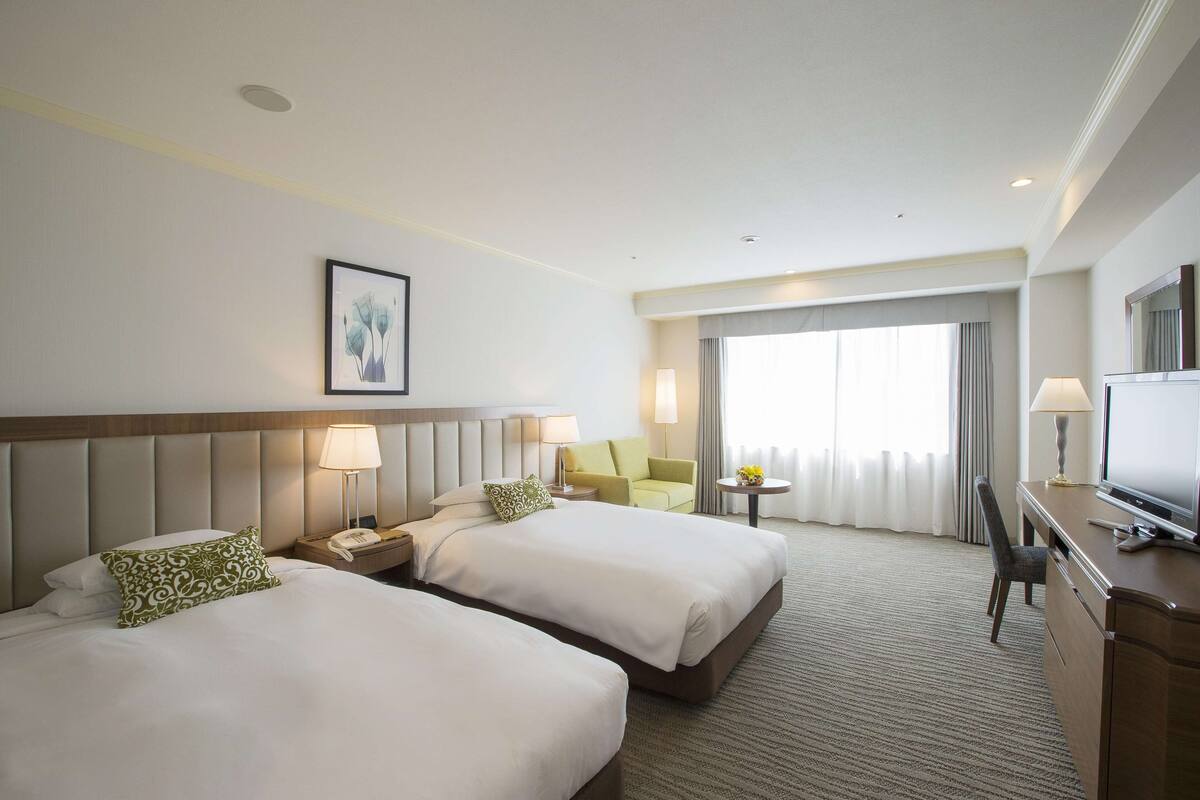 A hotel room with two beds, a desk, a chair, a television, and a window with curtains.