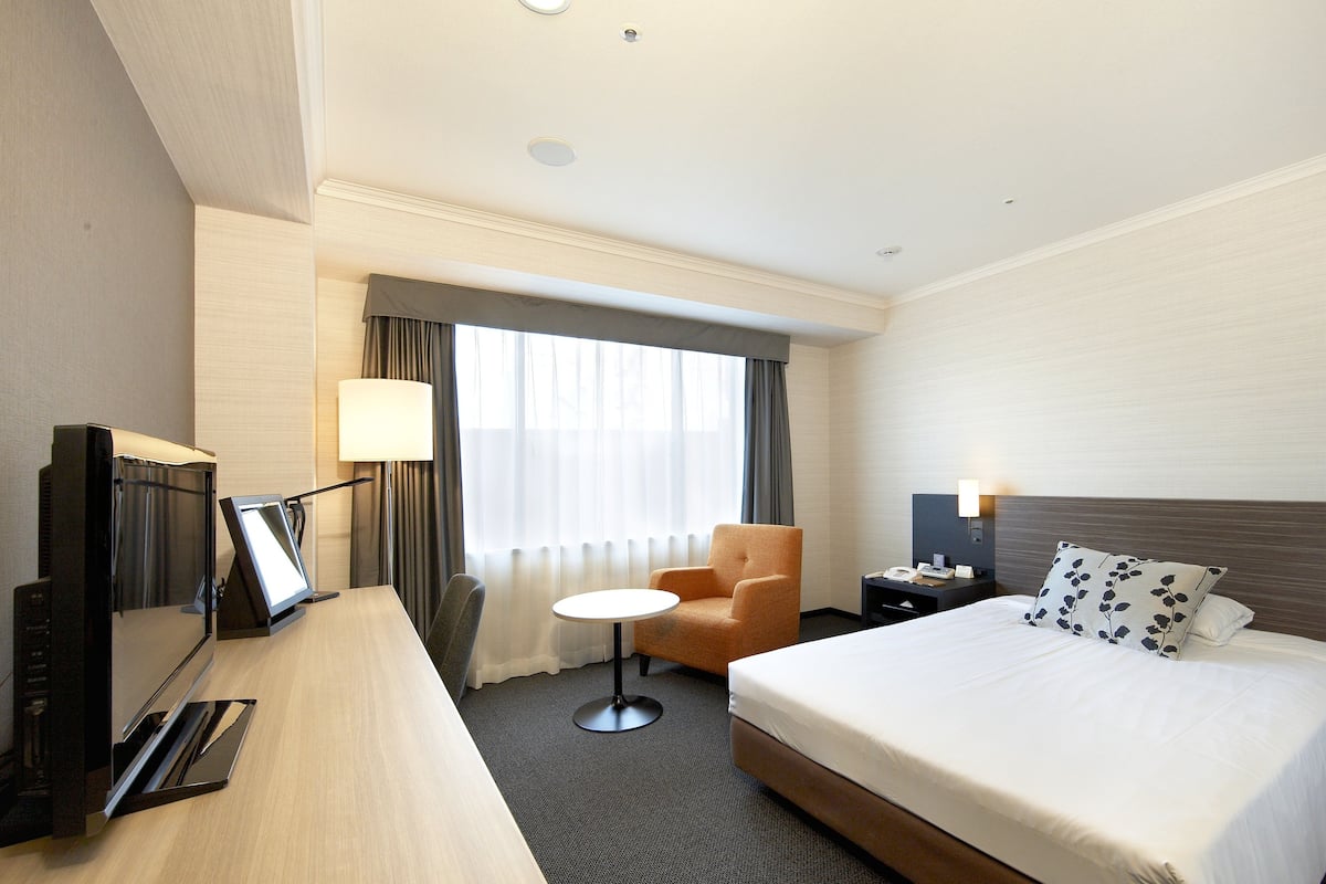 A hotel room with a bed, a desk with a computer, a chair, a small table, and a window with curtains.