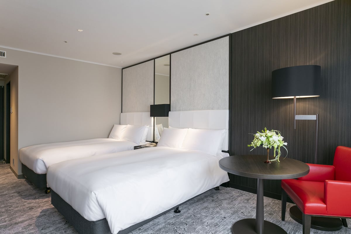 A hotel room with two beds, a round table, a red chair, a large mirror, and a wall-mounted lamp.