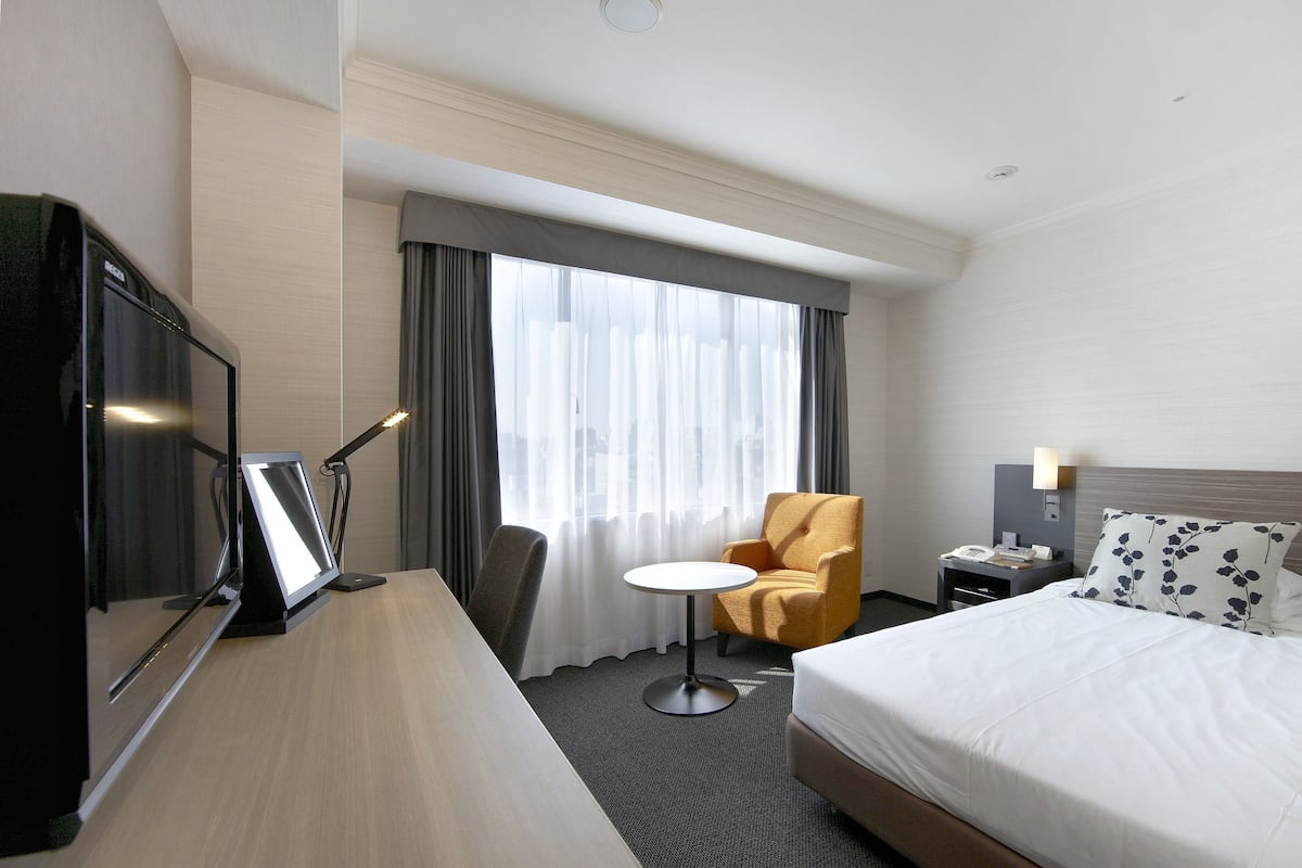 A hotel room with a bed, a desk with a computer, a chair, a small table, and a window with curtains.