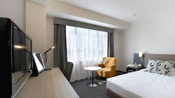 A hotel room with a bed, a desk with a computer, a chair, a small table, and a window with curtains.