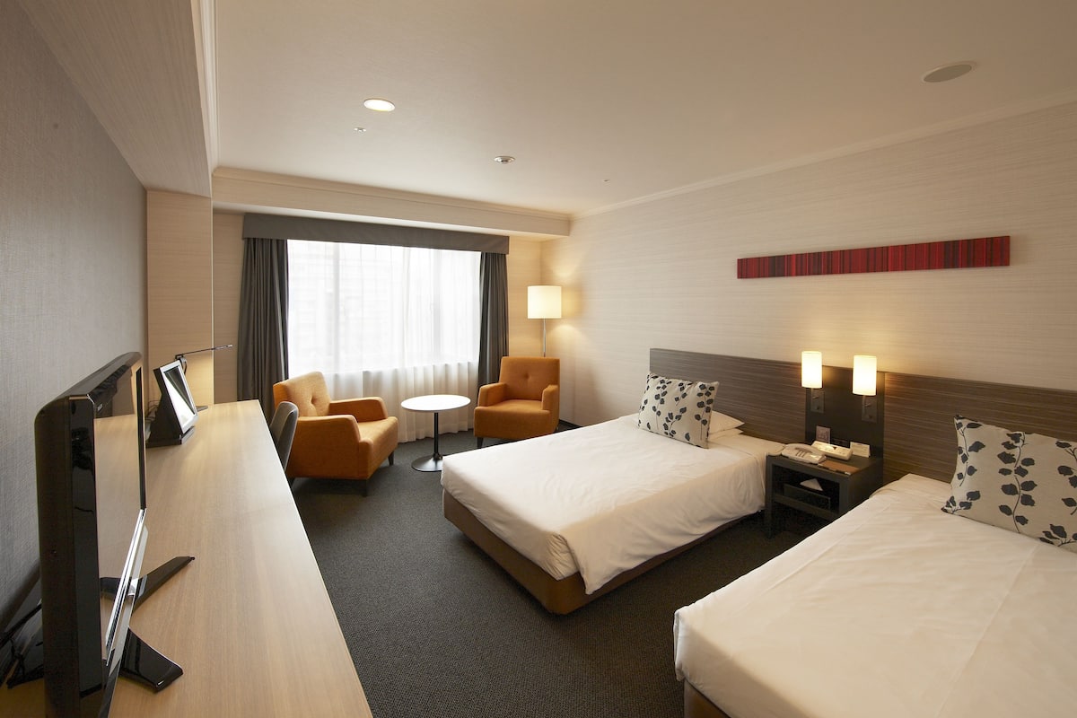 A hotel room with a large bed, a smaller bed, a desk with a computer, a chair, a lamp, and a window with curtains.