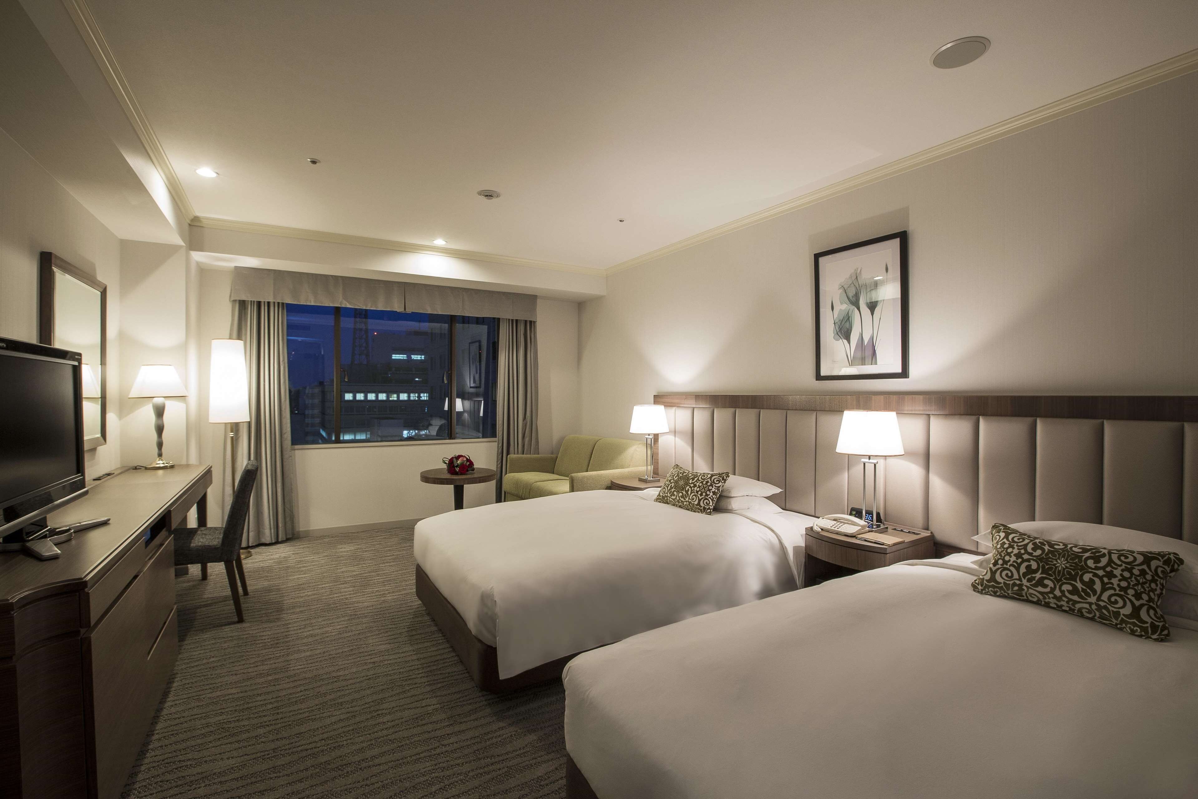 A hotel room with two beds, a desk with a TV, a chair, a small table, and a window with curtains.