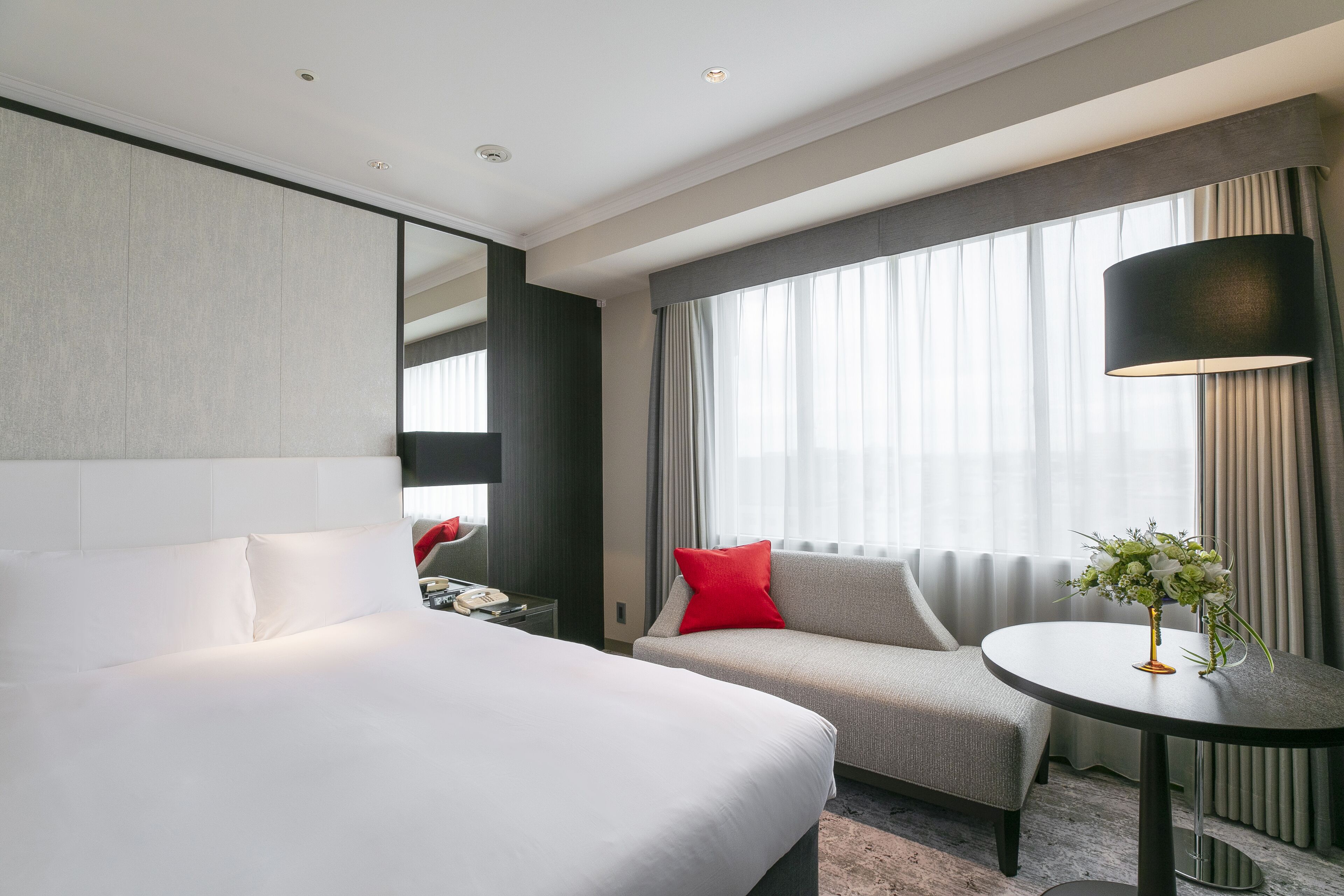 A hotel room with a large bed, a sofa, a small table with a vase, and a large window with curtains.