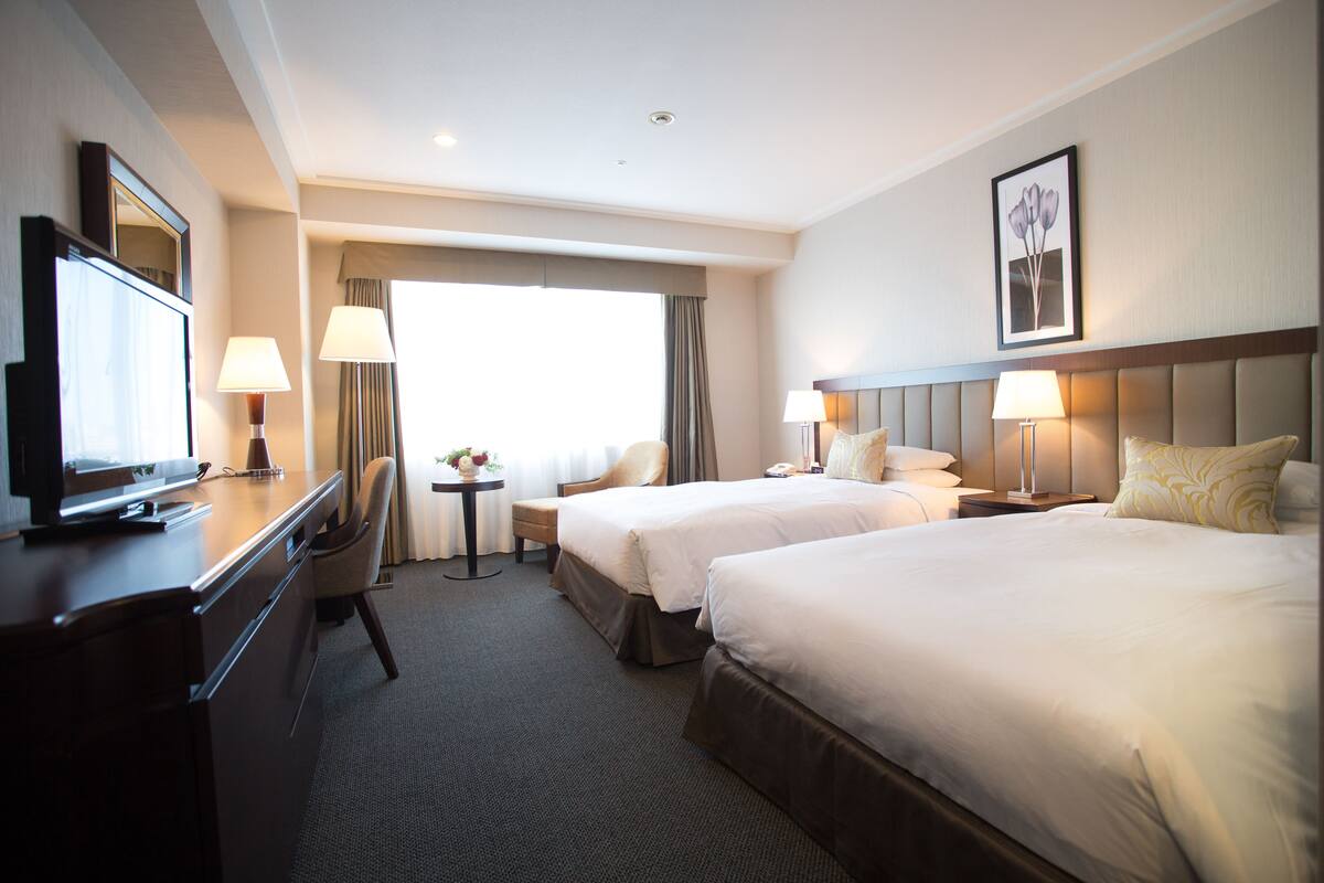 A hotel room with two beds, a desk with a TV, a chair, a small table with a lamp, and a window with curtains.