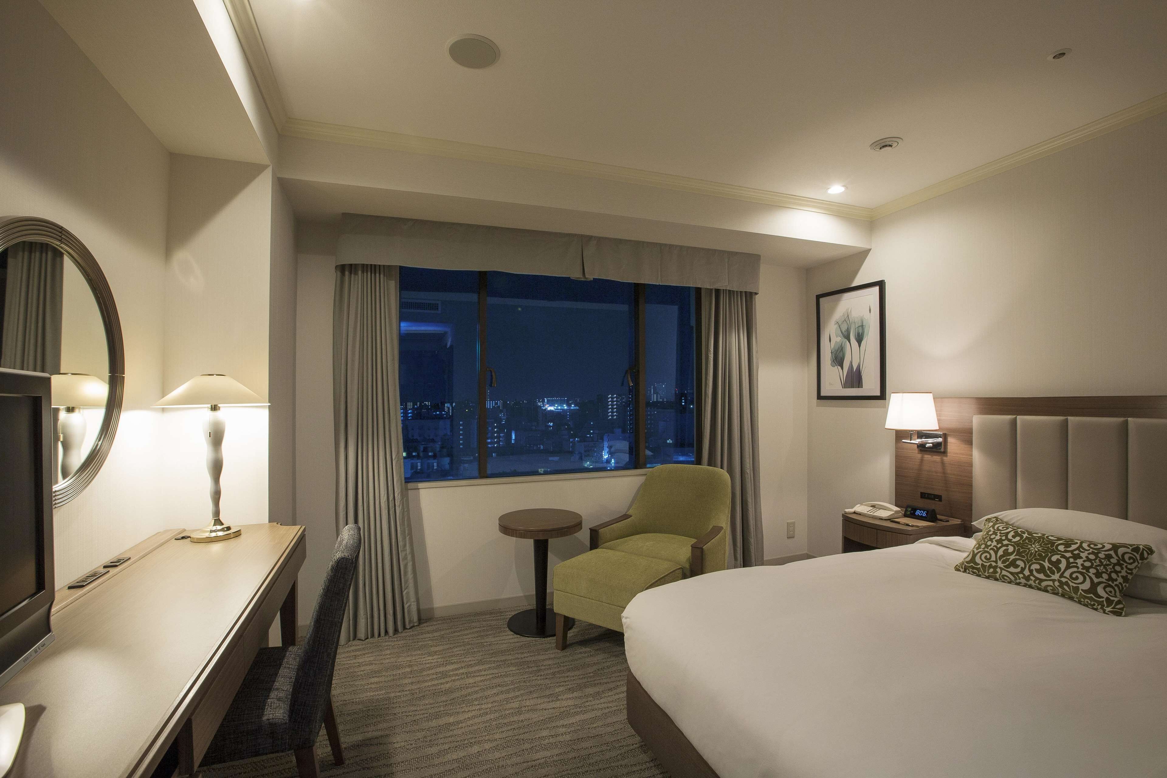 A hotel room with a large bed, a desk with a lamp, a chair, a small table, a nightstand with a phone, and a window with a city view.