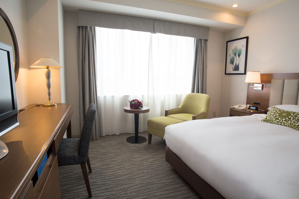 A hotel room with a bed, a desk, a chair, a small table with a lamp, and a window with curtains.