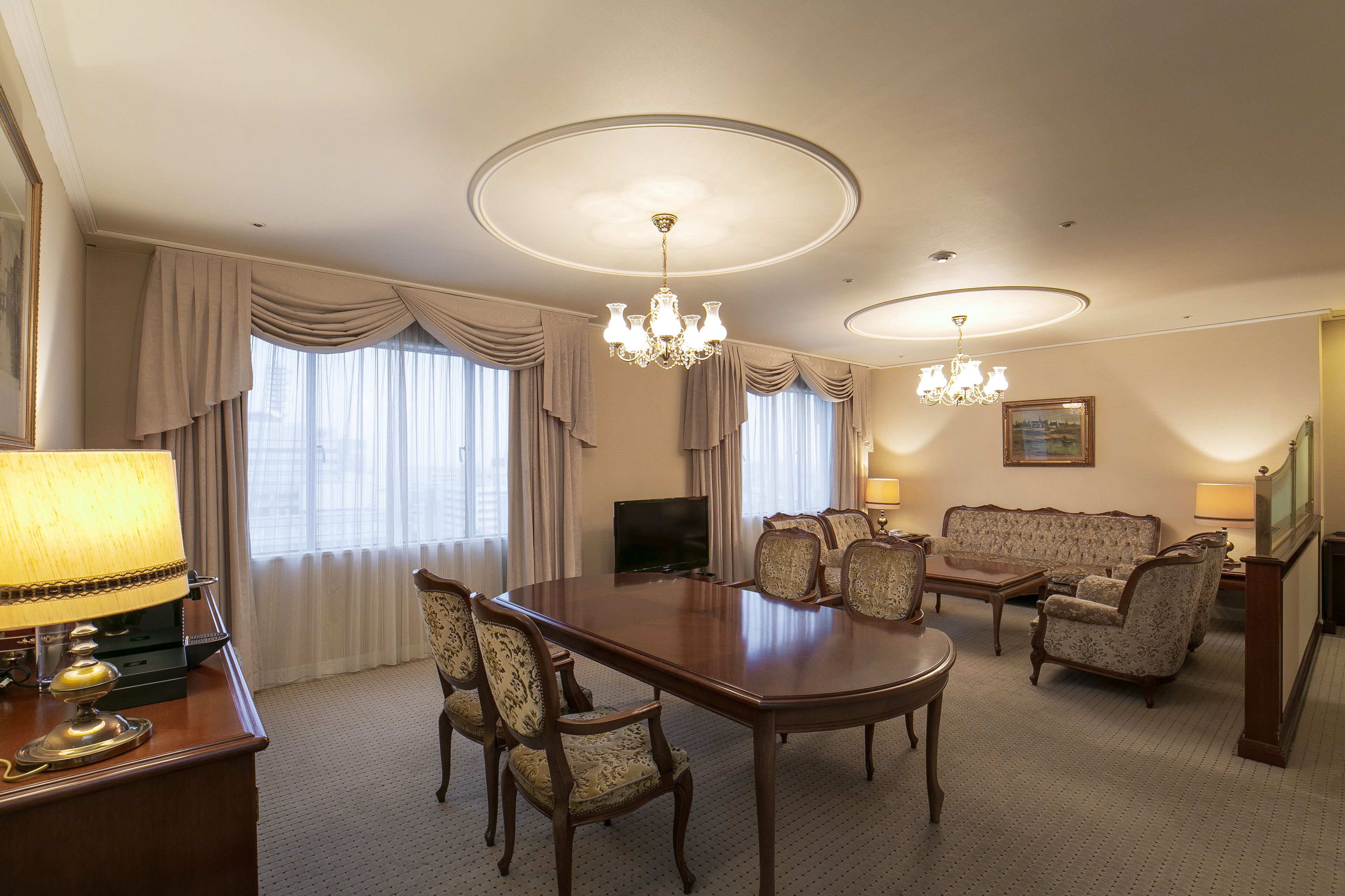 A hotel room with a dining table, chairs, a sofa, a TV, and a chandelier.