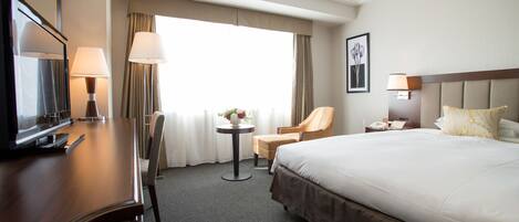 A hotel room with a large bed, a desk with a TV, a chair, a small table with a lamp, and a window with curtains.