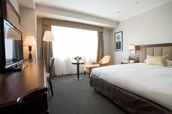 Habitación de hotel con una cama grande, un escritorio con televisor, una silla, una mesita con lámpara y una ventana con cortinas.