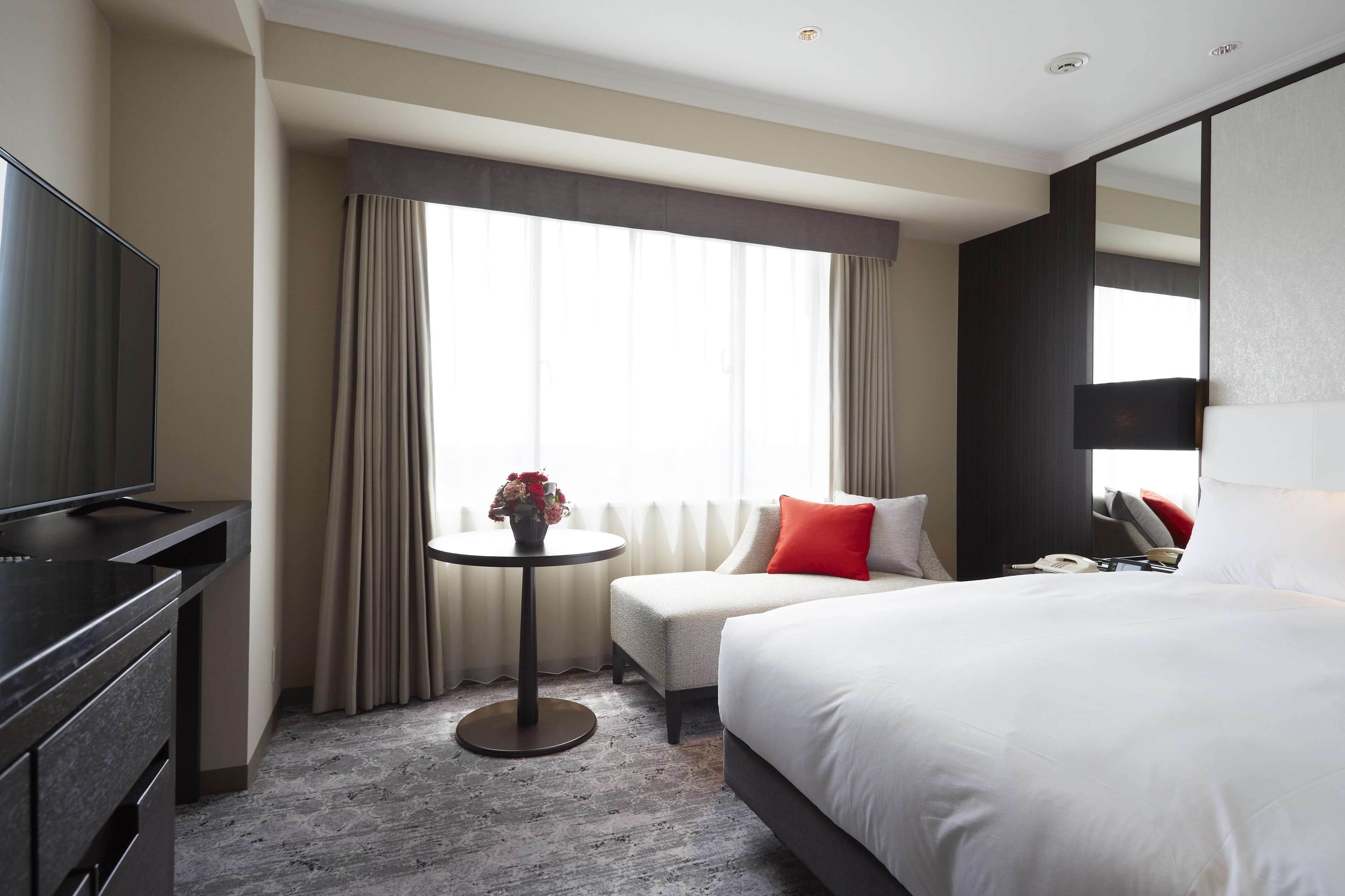 Habitación de hotel con cama, televisor, una mesita con un florero y ventana con cortinas.