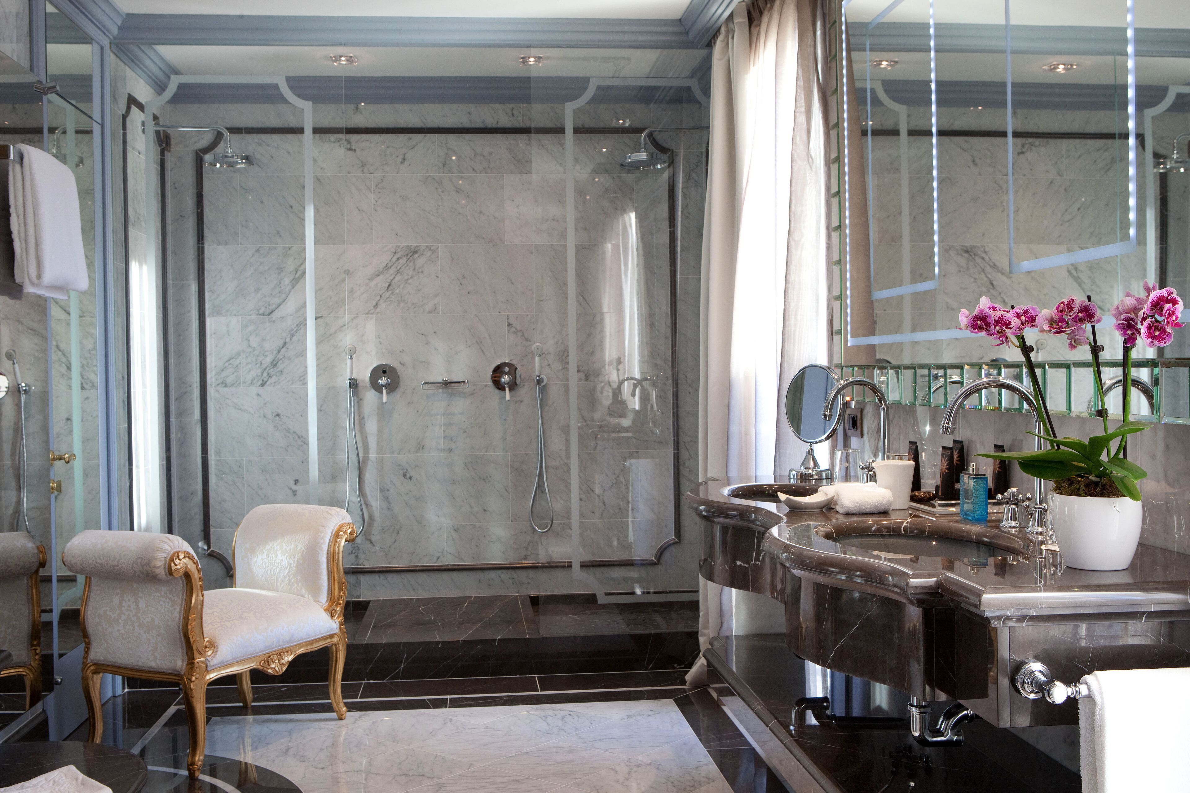 Une salle de bain luxueuse avec une douche en marbre, un fauteuil à cadre doré, un grand lavabo et une orchidée en pot.