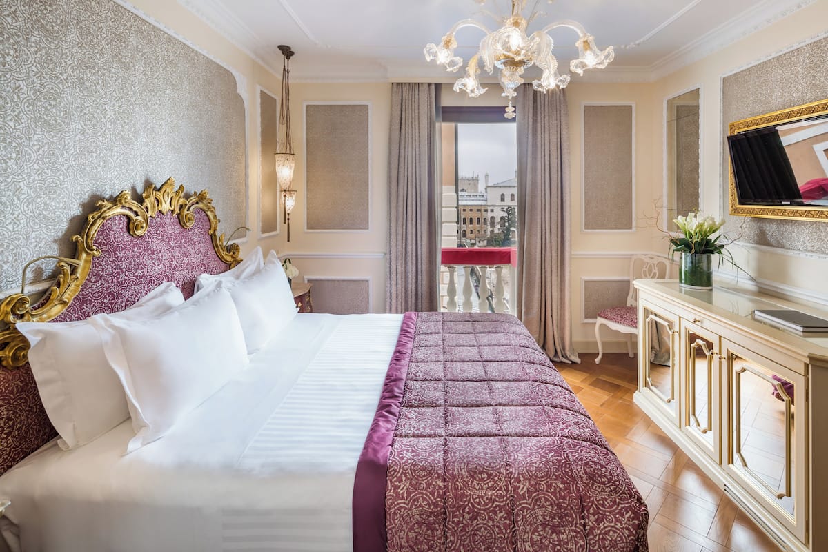 Hotellihuone, jossa on suuri sänky, kattokruunu, näkymä rakennuksiin ja televisio, joka on sijoitettu kaappiin.