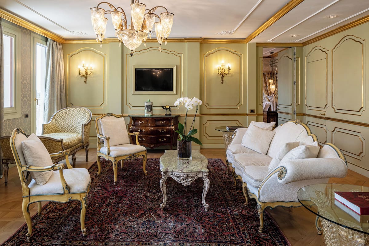 Un salon luxueux, agrémenté de meubles richement décorés, d’un lustre et d’un grand téléviseur à écran plat.