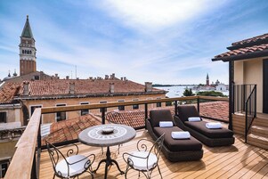 Terraço com mesa e duas espreguiçadeiras, com vista para um horizonte urbano com edifícios históricos e um rio.