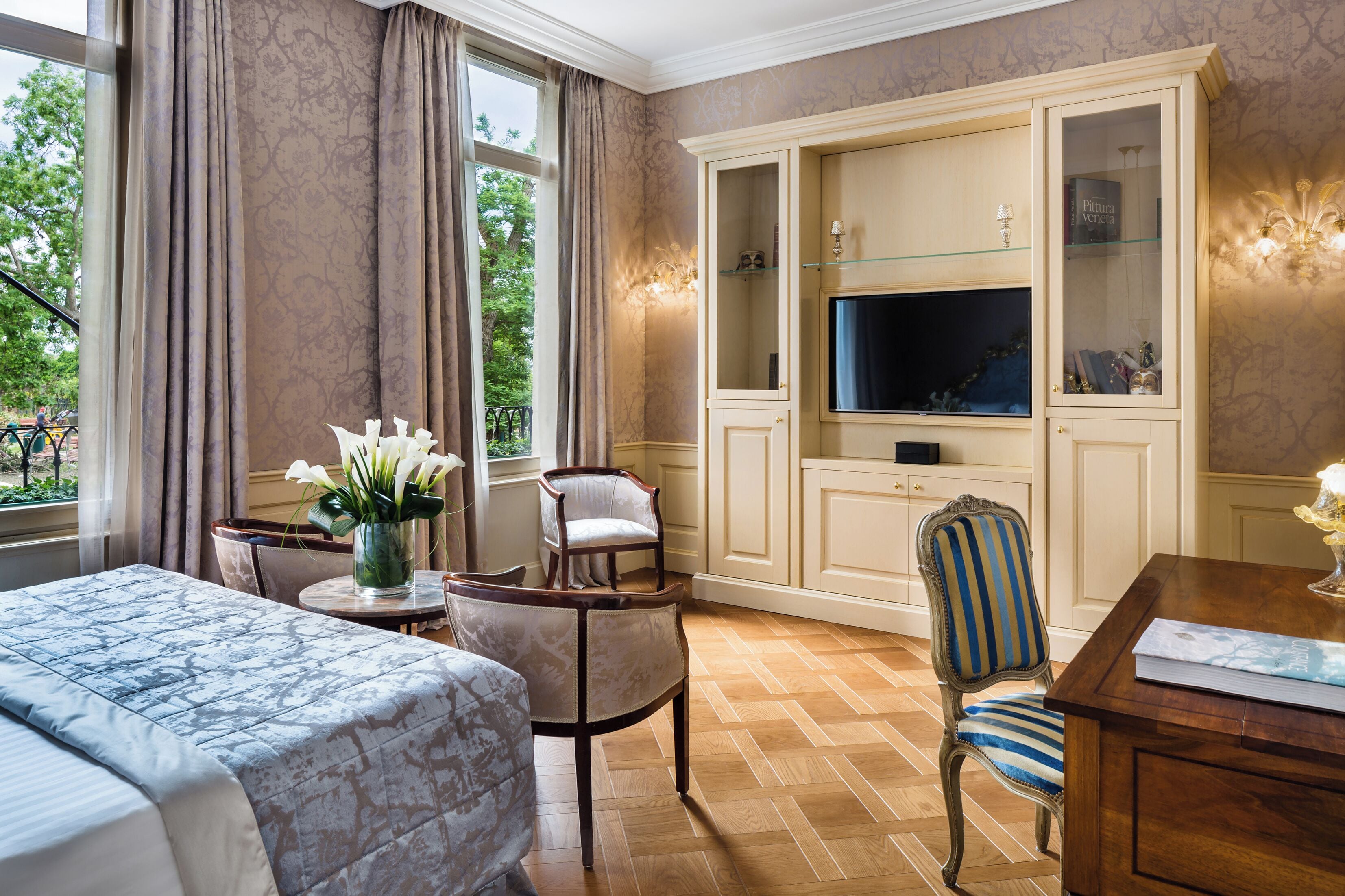 A hotel room with a bed, a TV, a seating area with a chair and a table, and a large cabinet with glass doors.