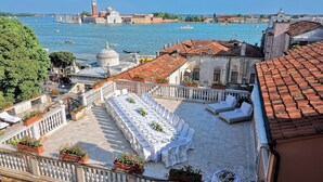 A terrace with a long dining table set for a meal, overlooking a canal with boats and historic buildings.