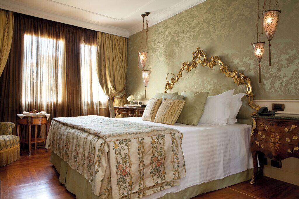 A large bed with a floral bedspread, a nightstand with a lamp, and a chair in a room with patterned wallpaper and curtains.